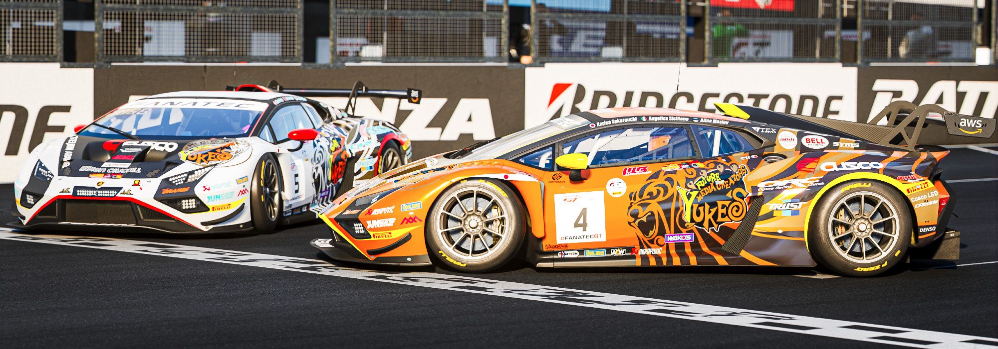 two Lamborghini Huracan GT3 Evo2 in front of a starting line; featuring a lion graphics holding the Yuke's logo in its claws and "showing" it to the viewer. one is orange with yellow accents and the other is white with red accents