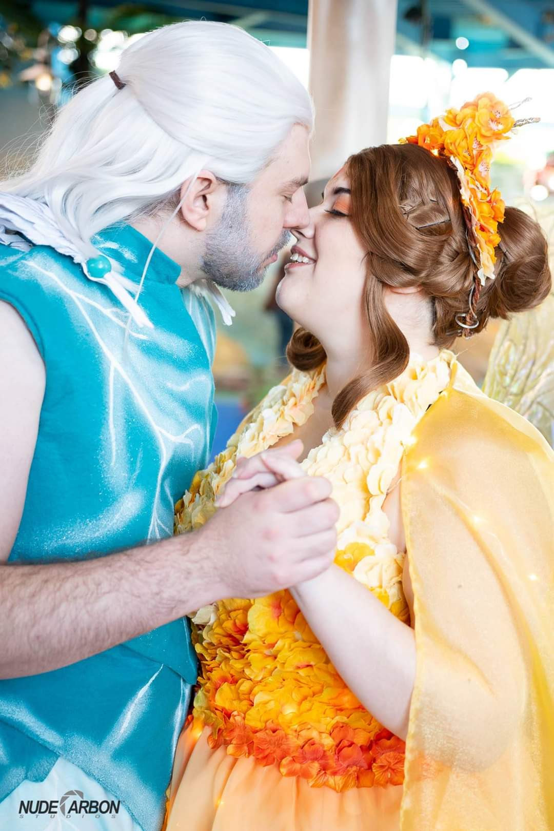 Two figures kissing.  The man in the photo has white hair and a blue-green vest that appears to be made of frosted leaves.  He has a cloak of owl feathers.  The woman is covered head to toe in hydrangea petals that color gradient from deep orange to pale yellow.  She has golden wings and a crown.