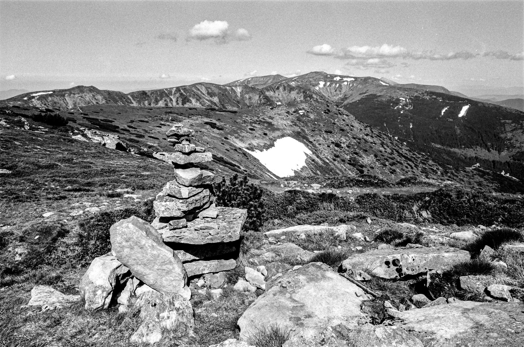 This black and white image depicts a mountainous landscape. In the foreground, there is a cairn, a pile of rocks carefully stacked on top of each other. The terrain is rocky and covered with sparse grass, while the background shows rugged mountain slopes with patches of snow remaining in some areas, especially visible on the right side. The mountains are rolling and extend into the distance, with ridges and valleys creating a sense of depth. The sky is clear, with a few small clouds scattered above the mountains, contributing to the serene and expansive feeling of the scene.