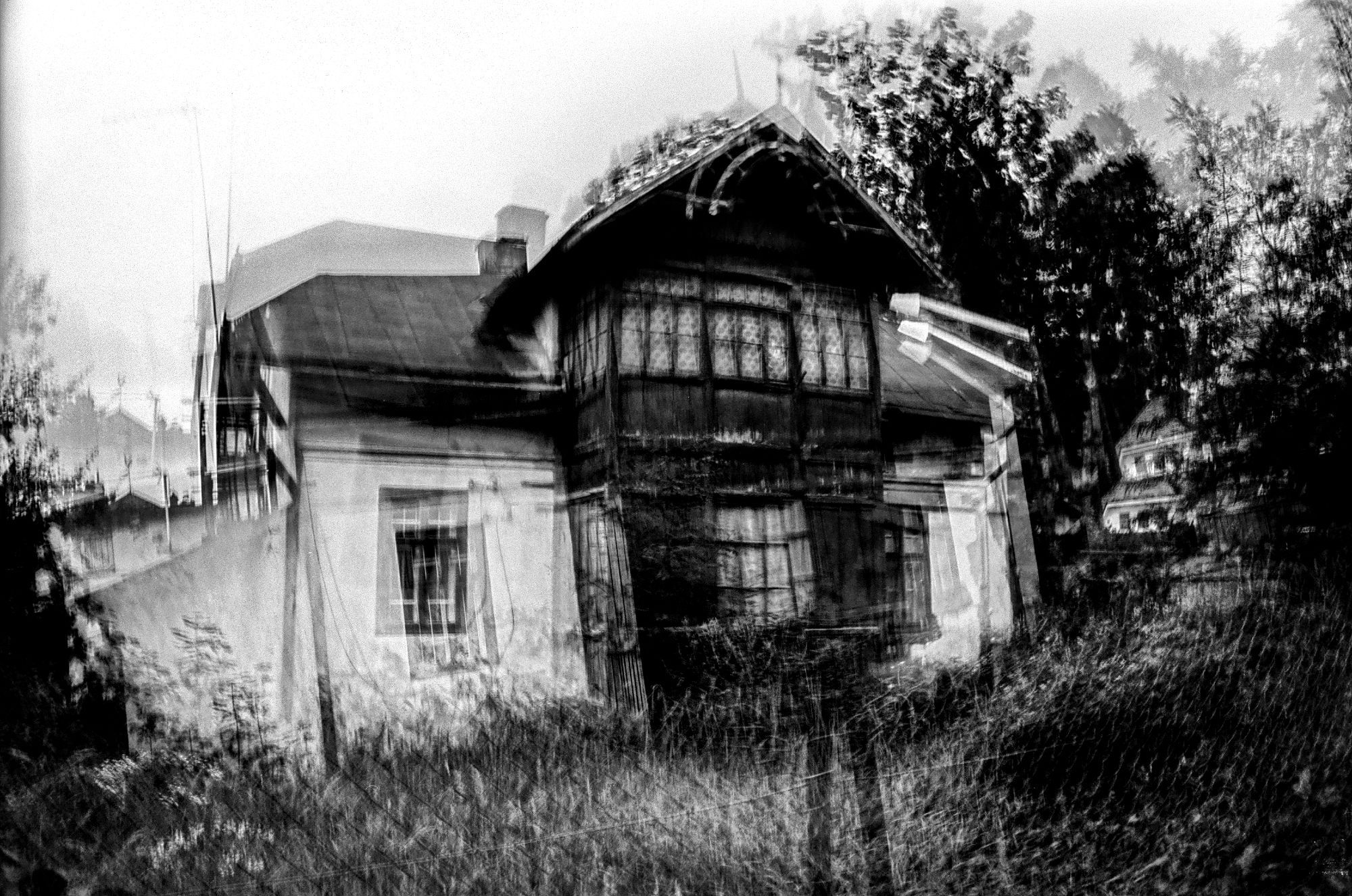 This image is a black-and-white photograph with a surreal, blurred effect that creates a ghostly, distorted appearance. The focal point of the image is an old, weathered house, which appears to be partially obscured by overgrown vegetation. The house has a steep, slanted roof and a large, central window area, possibly a bay window, with intricate wooden details. The overall scene is somewhat disorienting due to the double exposure or motion blur, making the house and surrounding trees appear as though they are shifting or melting into one another. The photograph has a haunting, eerie quality, as if capturing the remnants of a forgotten or decaying place.