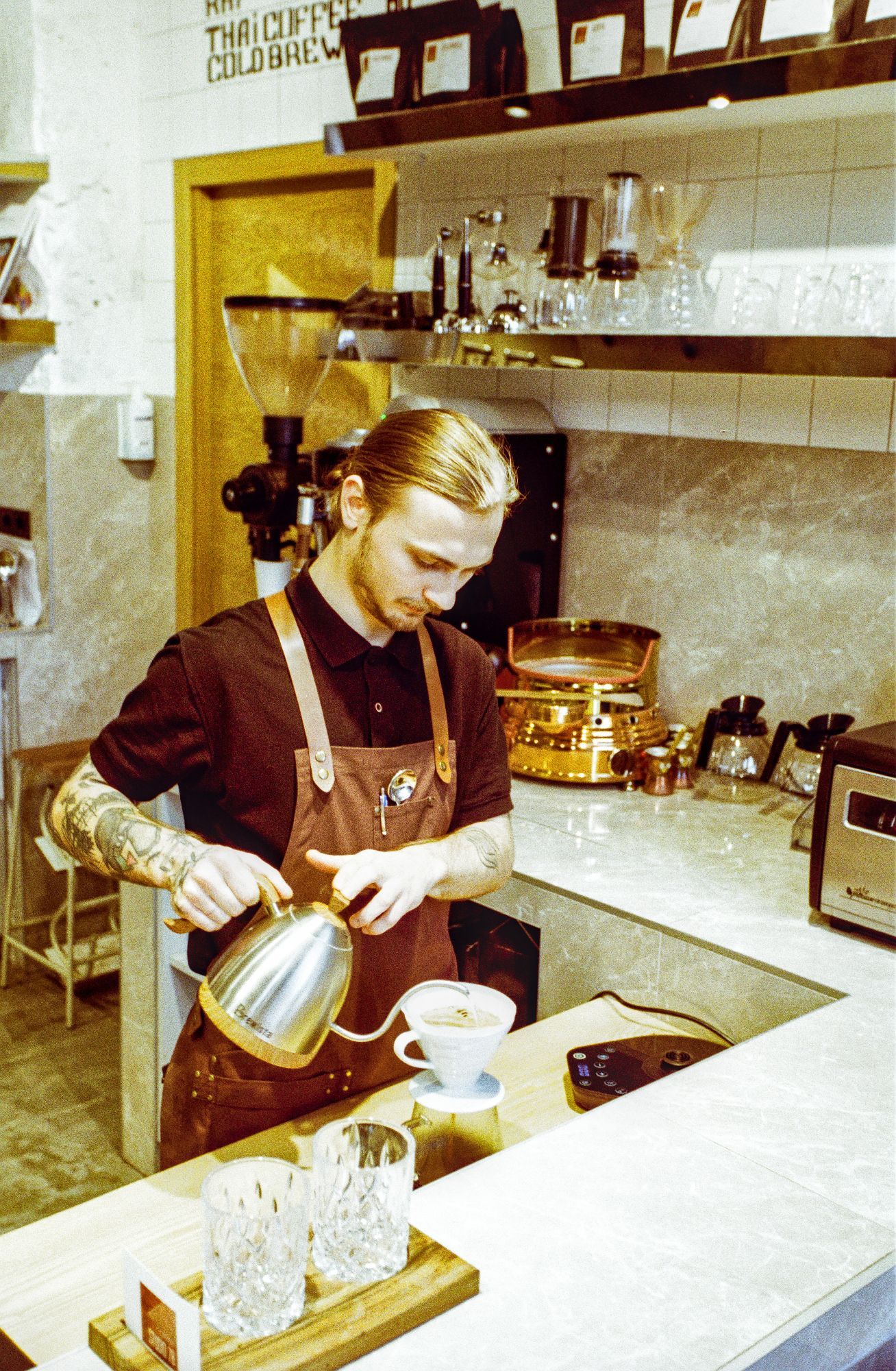 The image shows a barista with light-colored hair tied back in a bun, wearing a brown apron over a black shirt. He is carefully pouring hot water from a stainless steel gooseneck kettle into a white pour-over coffee dripper. The barista has visible tattoos on his arms, and the setting appears to be a modern coffee shop. There are glass cups, a wooden tray, and other coffee-making equipment on the countertop. In the background, various coffee brewing devices and bags of coffee are displayed on shelves, and a golden machine is visible, adding to the artisanal feel of the space.