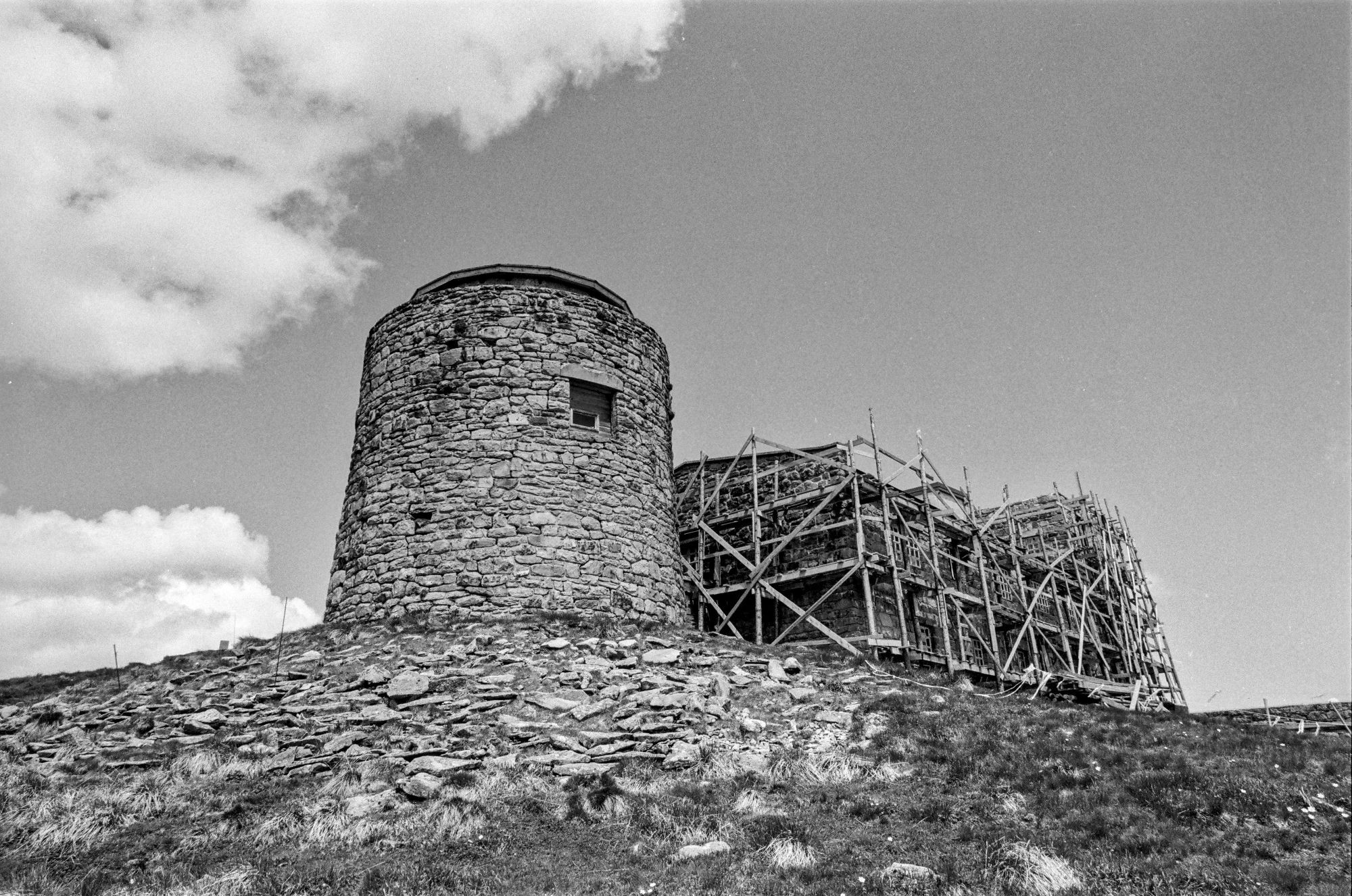 The image shows an old stone structure, which is part of the historic observatory located on Mount Pip Ivan, in the Chornohora Range of the Ukrainian Carpathians. The building appears to be under some restoration, with scaffolding surrounding part of it. The observatory is a cylindrical tower made of rough stone, with small windows and a sturdy, weathered appearance. The surrounding landscape is rocky and grassy, typical of a high-altitude environment. The black-and-white aesthetic of the photograph enhances the historic and remote atmosphere of this famous site, often referred to as the "White Elephant."