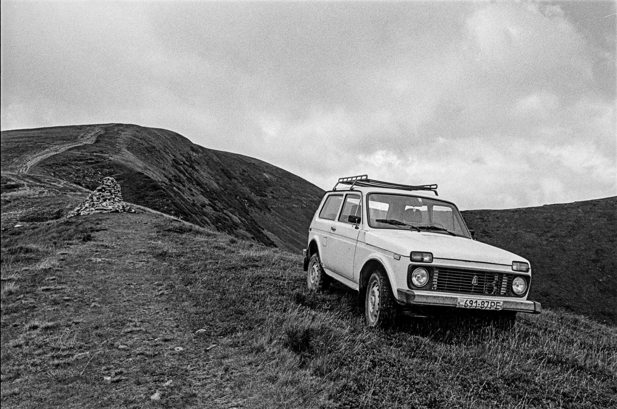 The black-and-white image depicts a rugged mountain landscape in the Ukrainian Carpathians. In the foreground, a classic Soviet-era off-road vehicle, a **Lada Niva**, is parked on a grassy hill. The vehicle is slightly tilted on uneven terrain, its license plate clearly visible. To the left, a stone cairn stands on the ridge, marking a trail that winds up toward a rounded, barren peak in the background. The terrain is wild and untamed, with no trees visible, just grassy slopes and rocky paths. Above, the sky is overcast, with thick clouds covering much of the horizon, adding a moody, atmospheric feel to the scene. The overall composition highlights the isolation and natural beauty of the remote mountains.