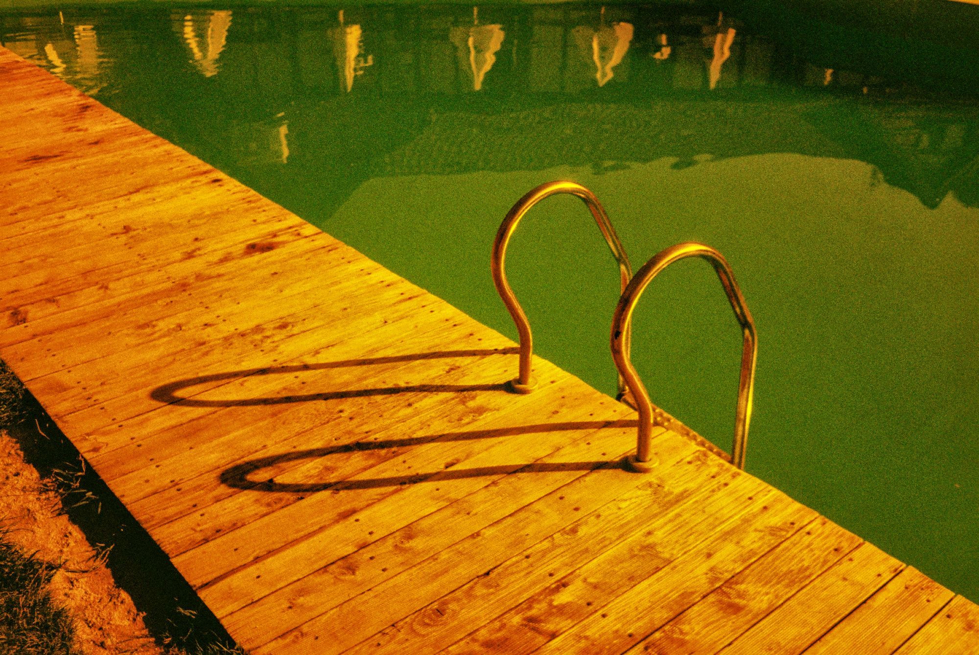 The image shows a wooden deck beside a swimming pool. The deck is lit with a warm, yellowish hue, casting deep shadows from the metal handrail that leads into the pool. The pool water has a greenish tint, giving a contrast to the golden tones of the deck. The reflection of nearby objects or lights is faintly visible on the water’s surface, adding a slight depth to the scene. The overall atmosphere feels calm and slightly surreal, with the lighting suggesting either early morning or late afternoon.