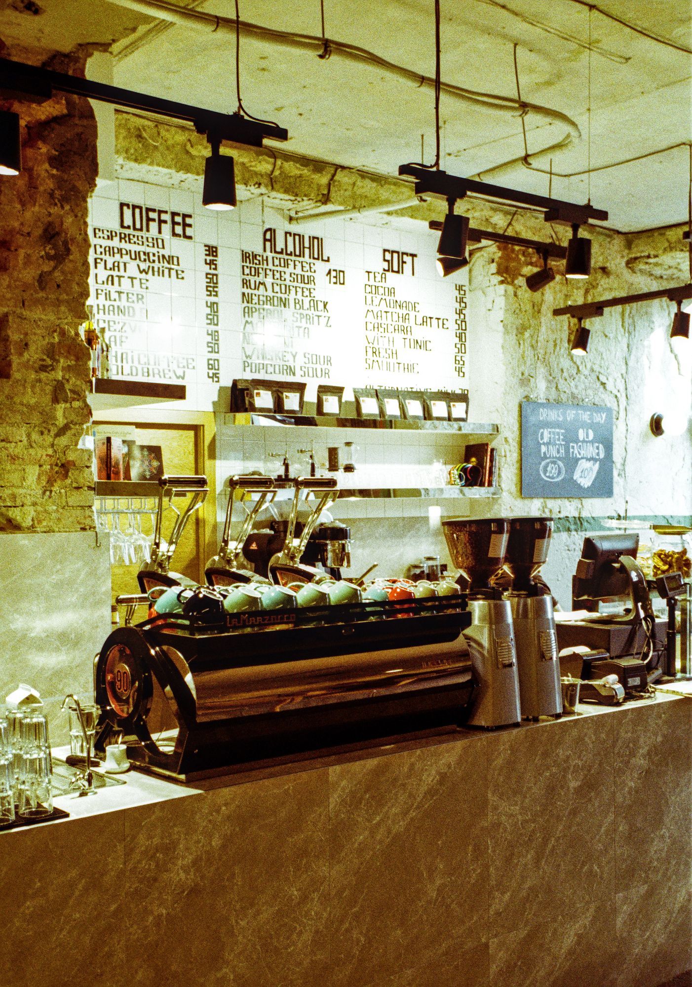 The image shows the interior of a coffee shop, focusing on the counter and menu board. The menu, written on white tiles, lists a variety of drinks, including coffee options like espresso, cappuccino, flat white, latte, and filter coffee. There are also alcoholic drinks, such as Irish coffee and Aperol Spritz, and soft beverages like tea, cocoa, lemonade, and matcha latte. The counter features a large "La Marzocco" espresso machine with colorful cups stacked on top, grinders, and other coffee-making equipment. The setting has an industrial and rustic aesthetic with exposed brick walls, hanging black pendant lights, and a chalkboard displaying the "Drinks of the Day." The overall atmosphere feels warm and inviting, blending modern coffee culture with a cozy, artisanal vibe.