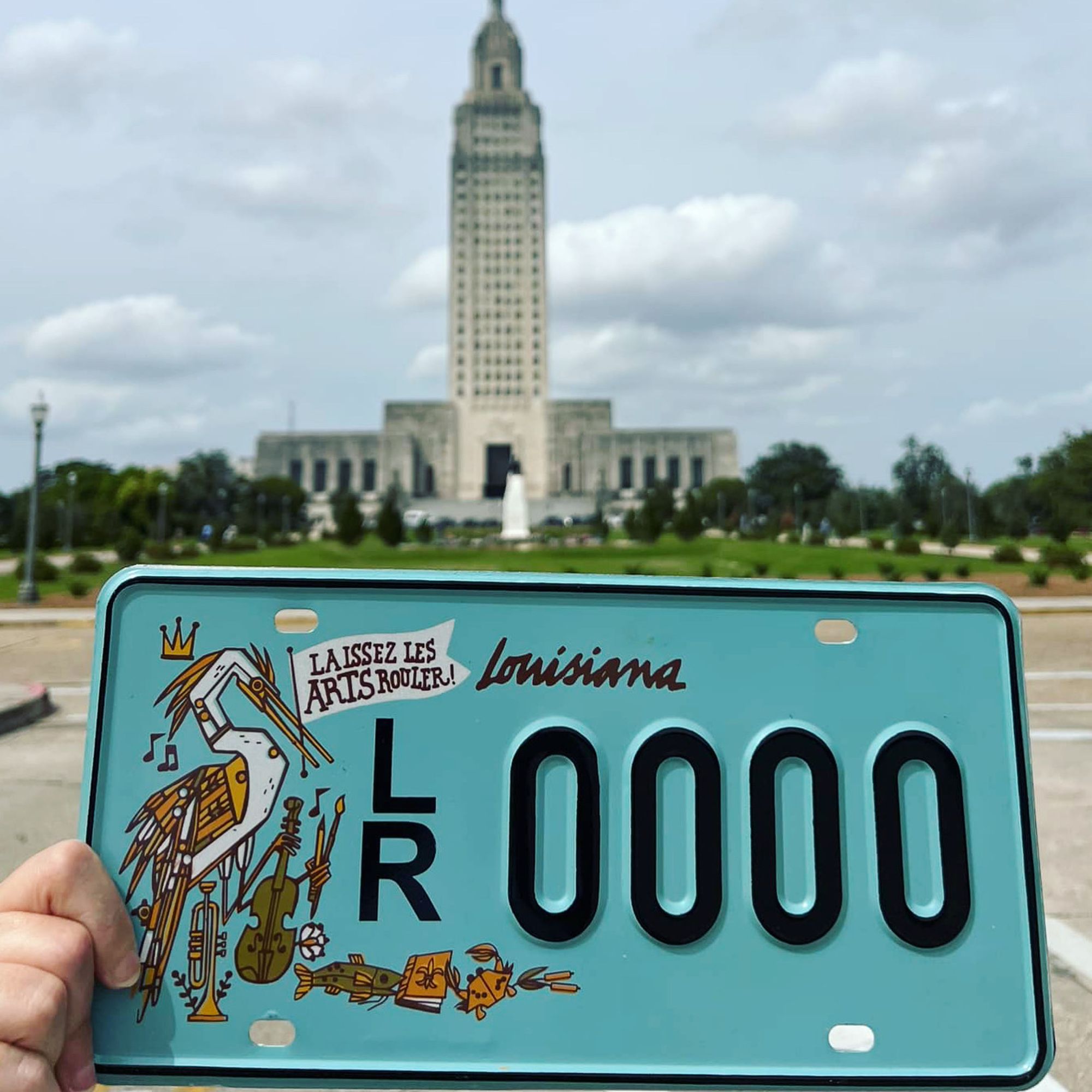 Photograph of the Louisiana Partnership for the Arts new license plate supporting the arts. The license plate is being held up with the State Capitol building in the background. The plate, designed by artist Denise Gallagher, is blue and features an egret holding a flag which reads "Laissez les Arts Rouler!" The bird is holding a violin, paintbrush and pencil. At its feet are a trumpet, catfish, book and crab. Each element representing the arts in Louisiana.