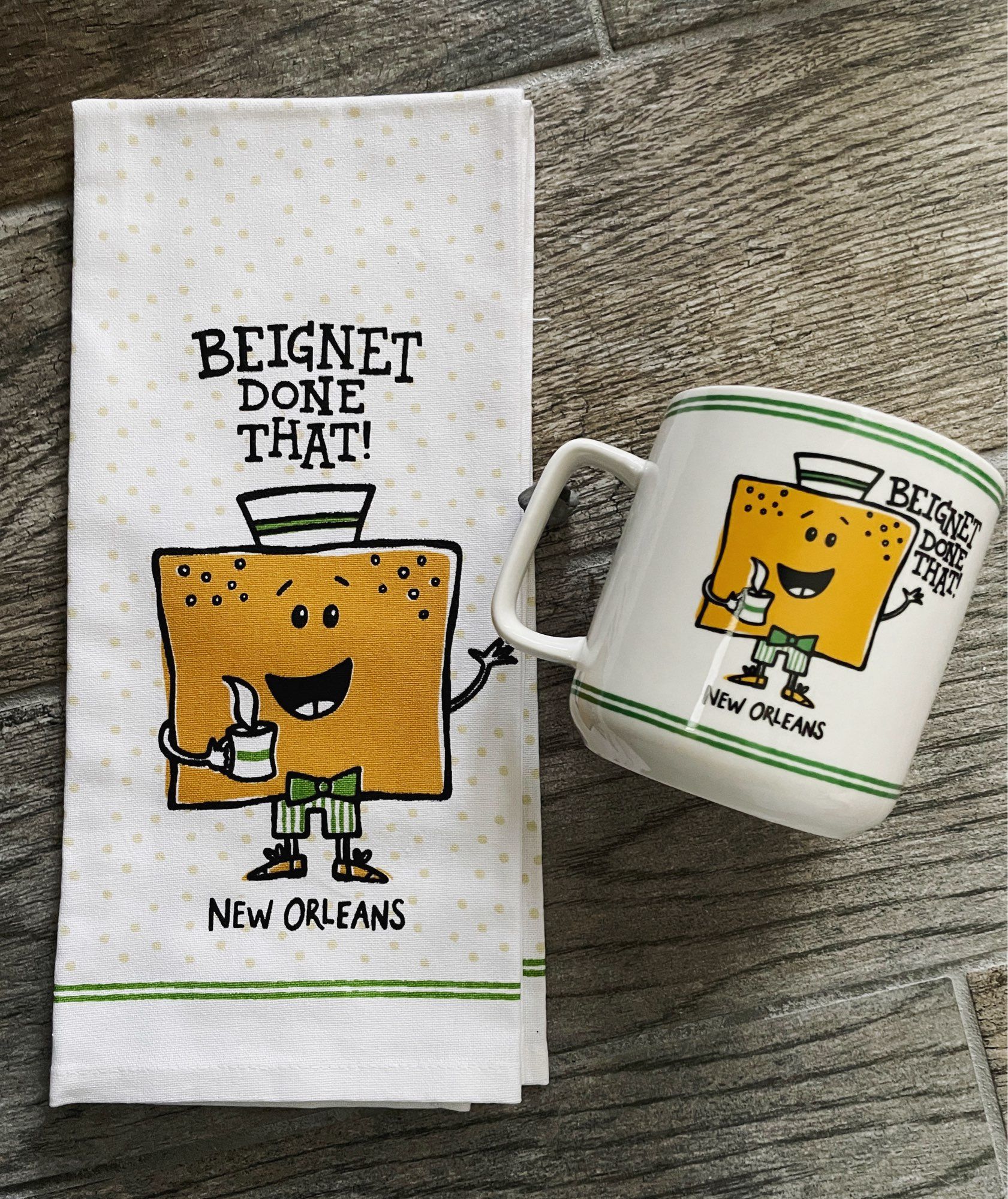 Photo of a kitchen towel and ceramic mug, both feature a design by artist, designer and illustrator Denise Gallagher. Hand lettered type reads “Beignet Done That!” above a beignet character with a paper hat, green bow tie, green striped pants and shoes. He holds a steaming cup of coffee and is waving.