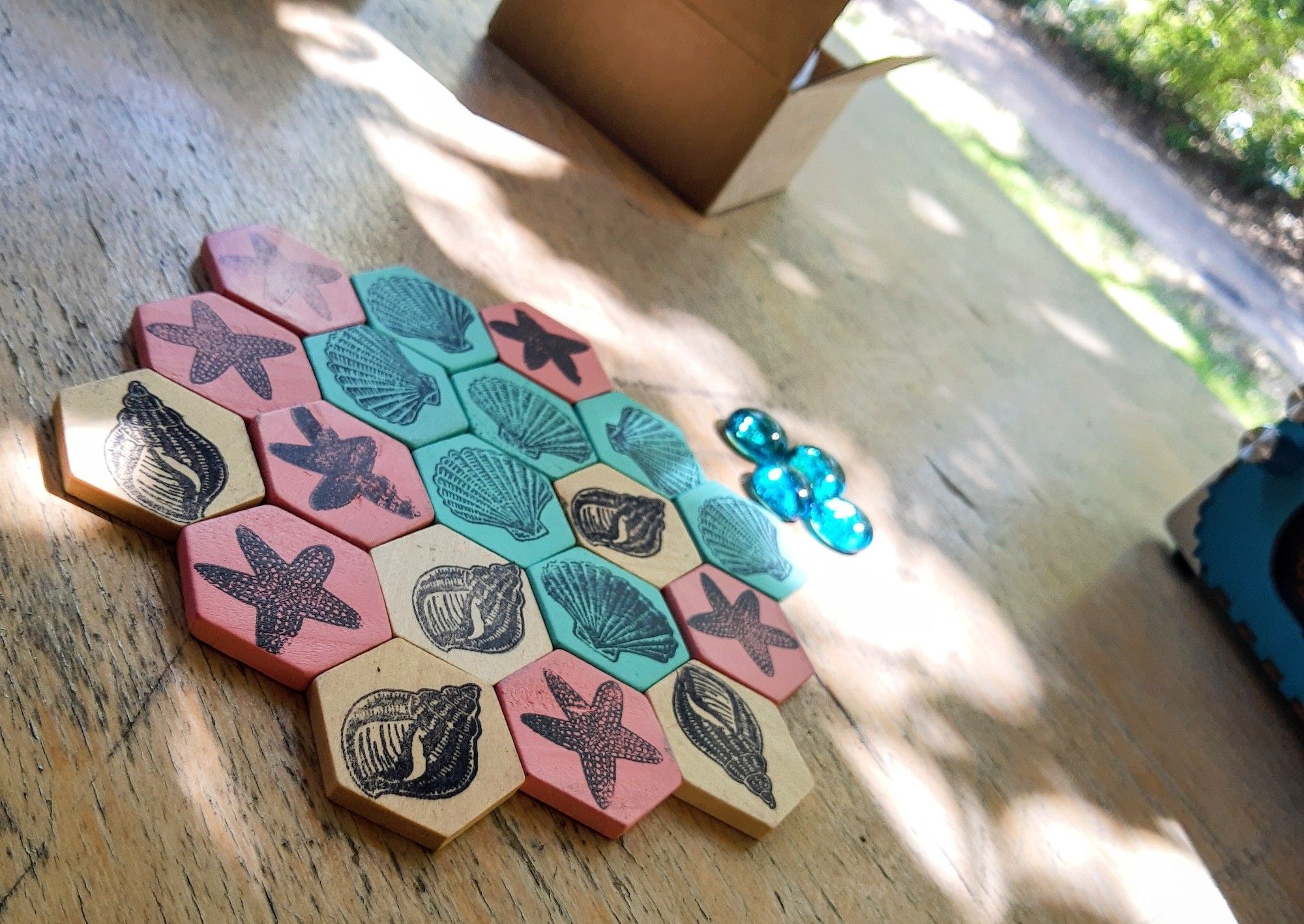 Hex tiles on a table, assembled into a larger hex (tiles in row of 3, 4, 5, 4, 3). The tiles are three different colors and labeled with conch shell, fan shell, and a starfish, and randomly placed in the formation. There are four glass counter stones.