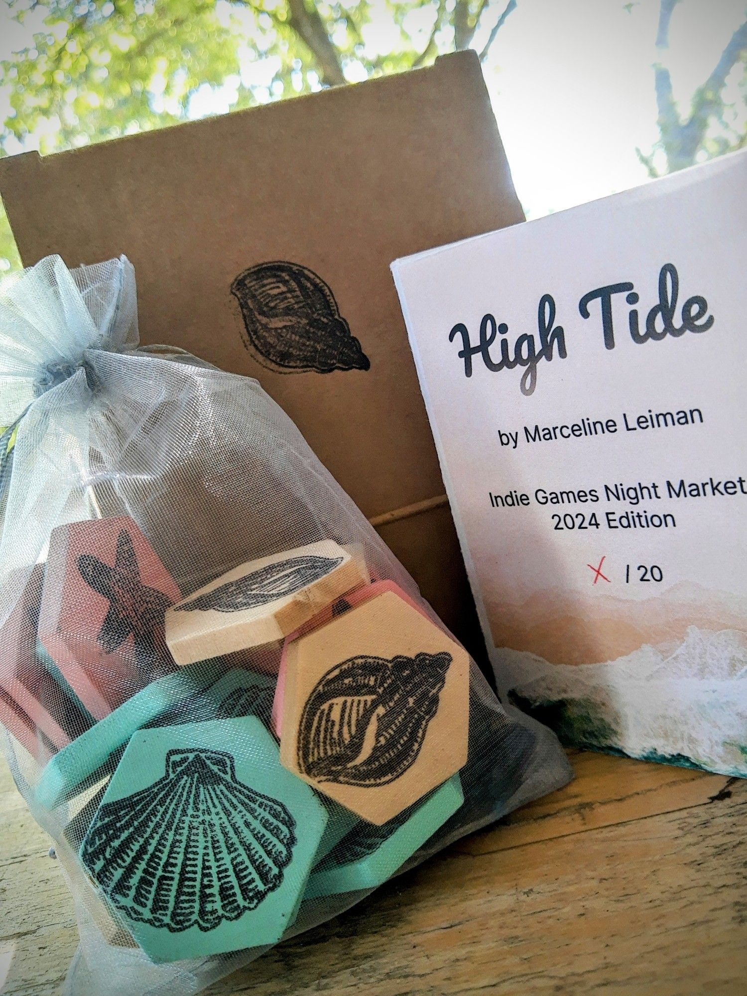 The colored tiles in a silver mesh bag, a cardboard box stamped with a shell, and a copy of the instruction booklet. Text on the booklet reads High Tide by Marceline leiman, Indie Games Night Market 2024 Edition, x/20.