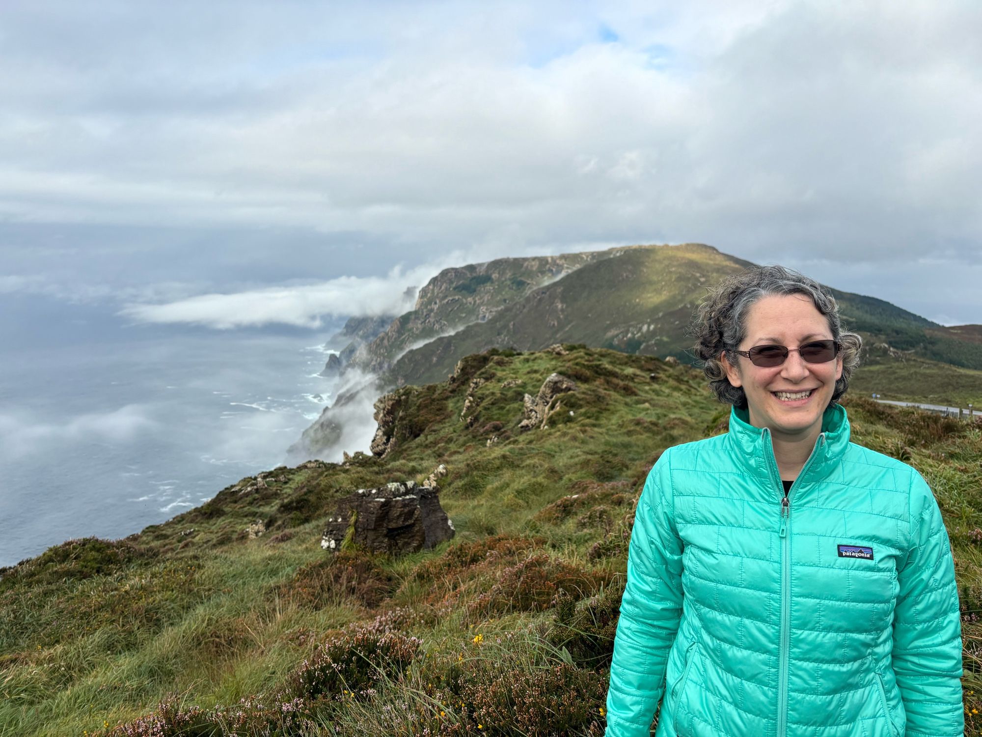 Woman in jacket stands in front of green rocky cliff by the ocean under cloudy sky