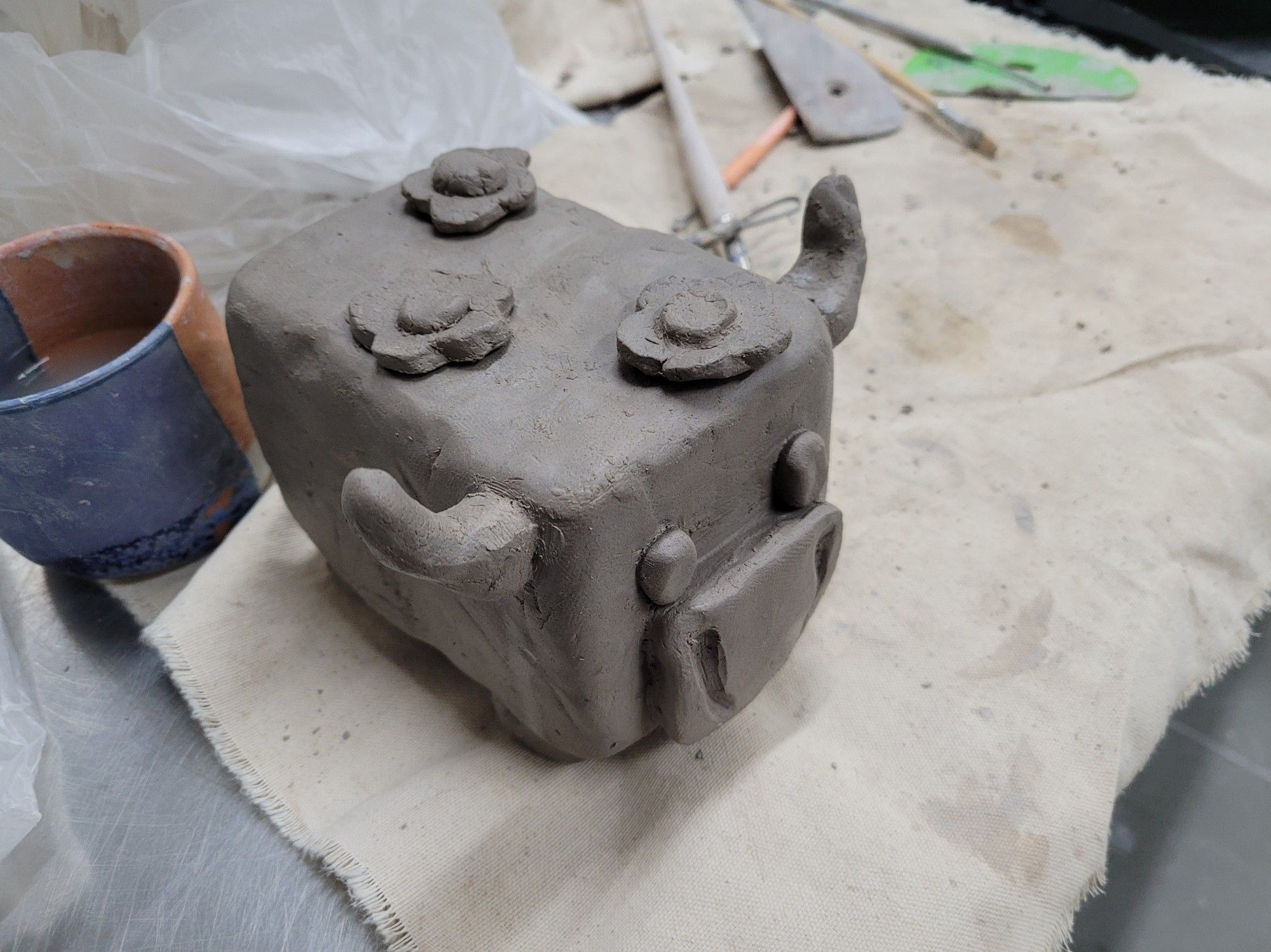 An in-progress ceramic cow. It is a blocky in shape, with very simplified features. Three small flowers grow out of its back. It's on a canvas with a pot of water on the side.