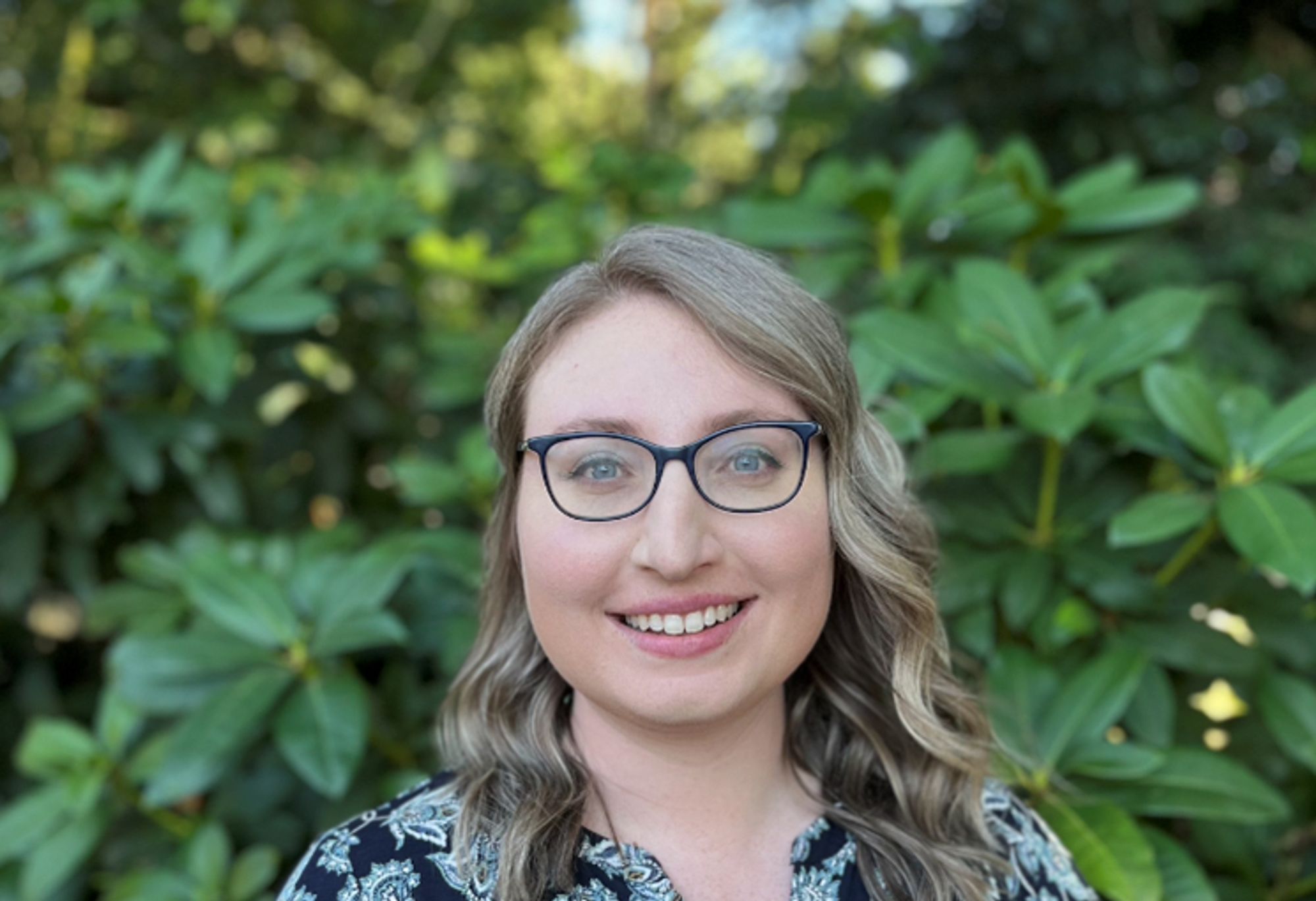 A woman with light skin and shoulder-length blond hair is smiling at the camera. She is wearing glasses with a rectangular blue frame and a black shirt with a floral pattern. The background consists of lush greenery, possibly a garden or a park, with soft sunlight filtering through the foliage.