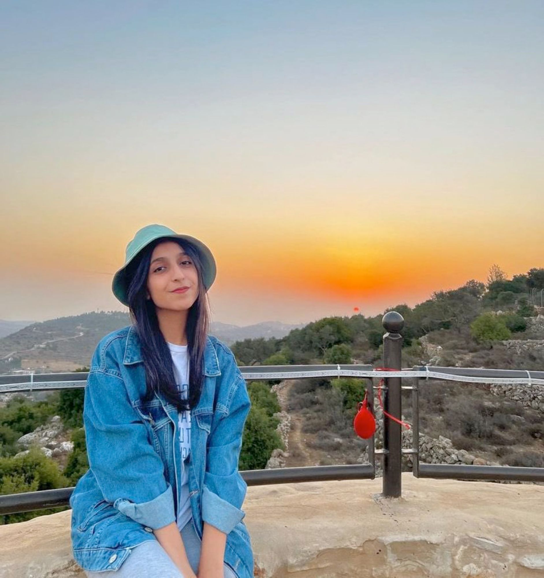 Photo of a young woman, university student Dua Al Qadi, taken at sunset high in the hills. She is wearing a t-shirt and jeans, denim jacket and denim sun hat, and smiling at the camera.