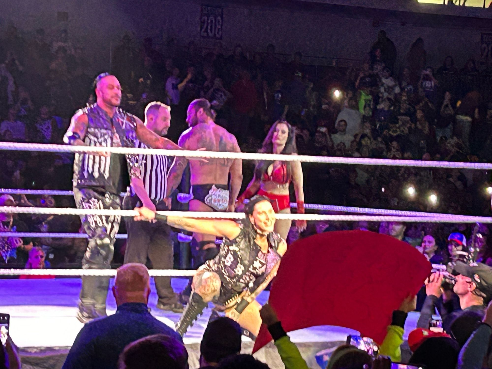 Wrestler Rhea Ripley hangs off the ring ropes making her match entrance. Her tag partner Damien Priest and their opponents are behind her