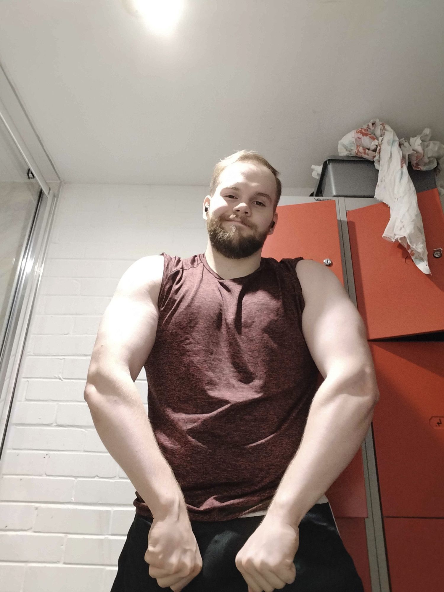 A man in a red shirt flexing his arms inward to show off his forearm definition while smirking at the camera.