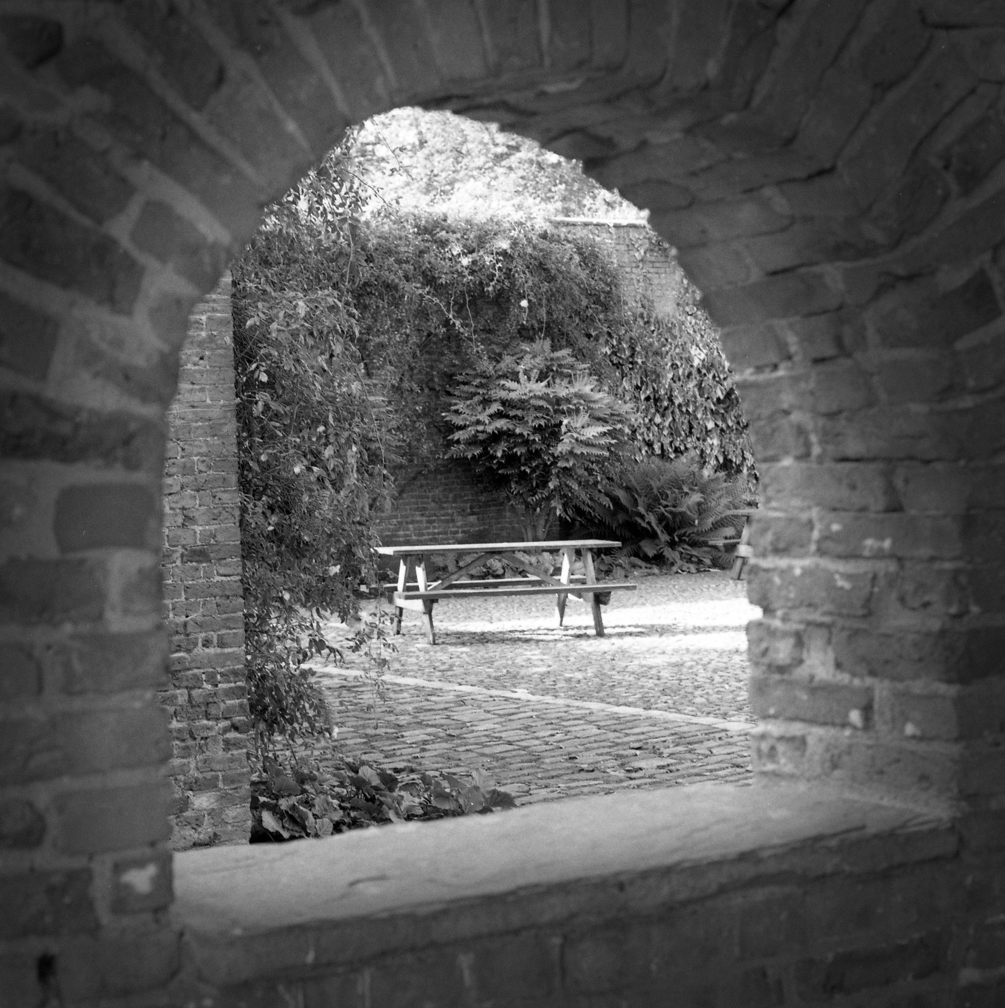 A black and white photograph shot through an arched opening, showing a picnic bench in a sunny courtyard