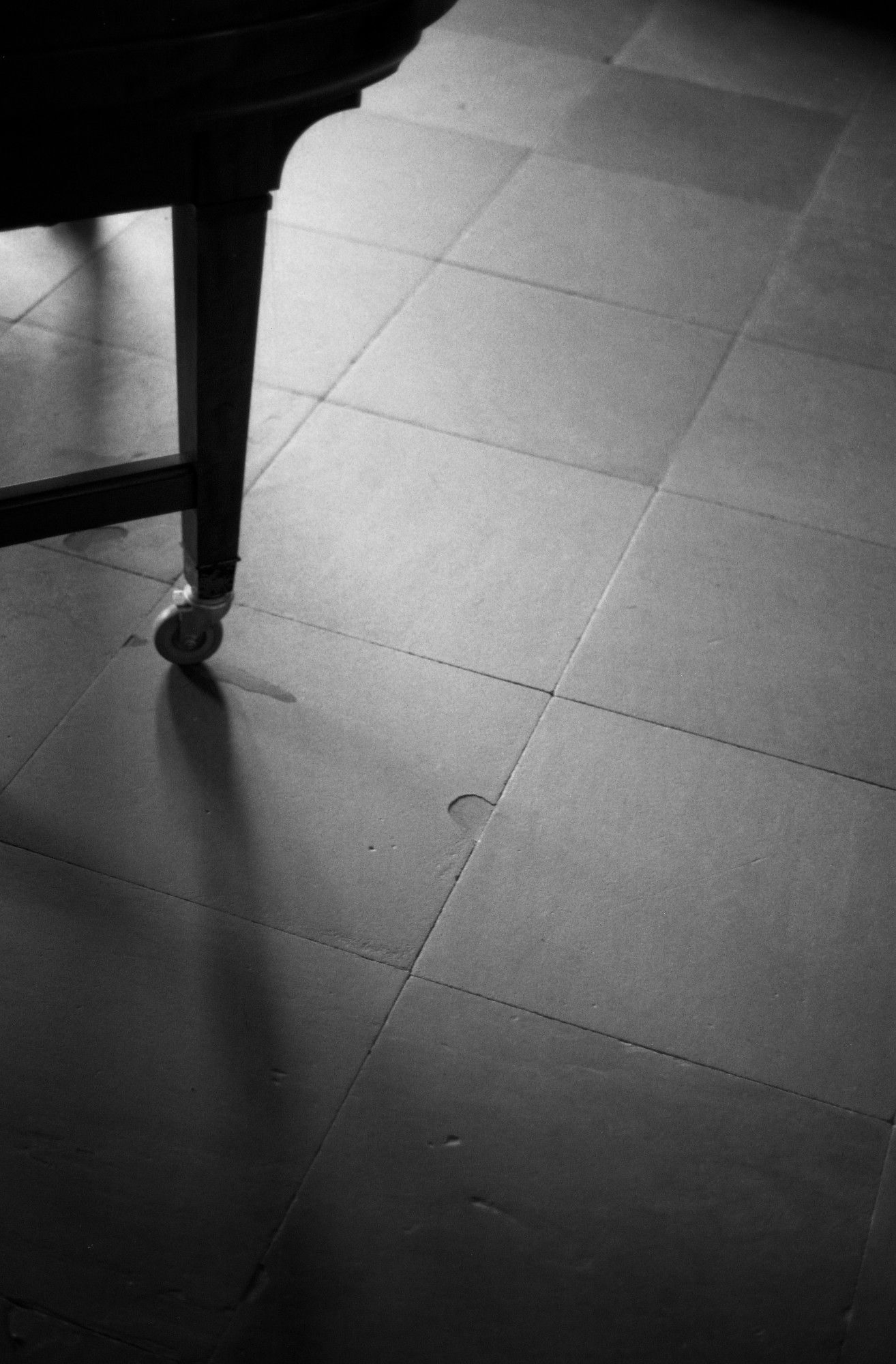 A black and white photograph showing a corner of a piano and its shadows on a stone floor