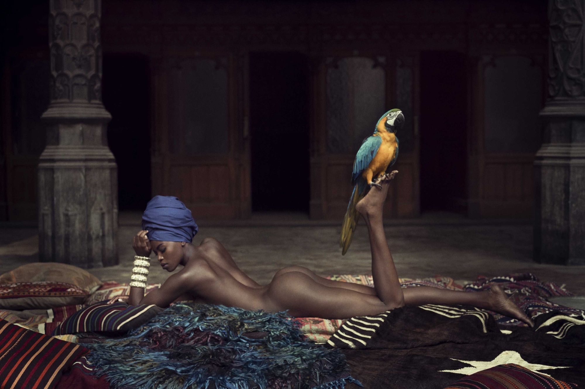 a beautiful naked brown lady laying facedown on a rug, in an Eastern palace, with her left foot turned up and a blue and orange macaw perched on it