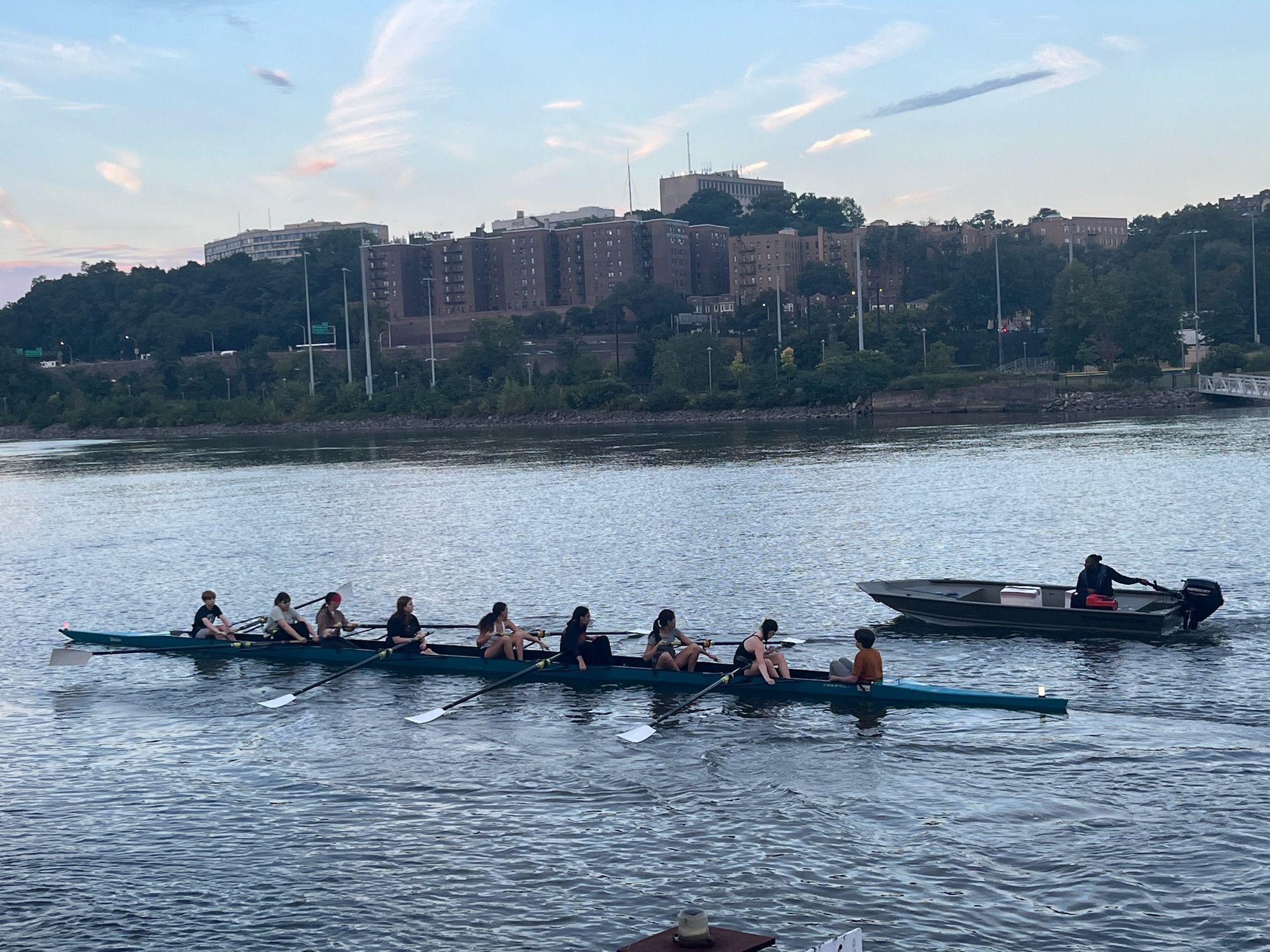 8 person row boat with coxswain pause on a river glittering under the sky at dusk with chase/support boat near by. Large buildings of TheBronx nyc in the background.