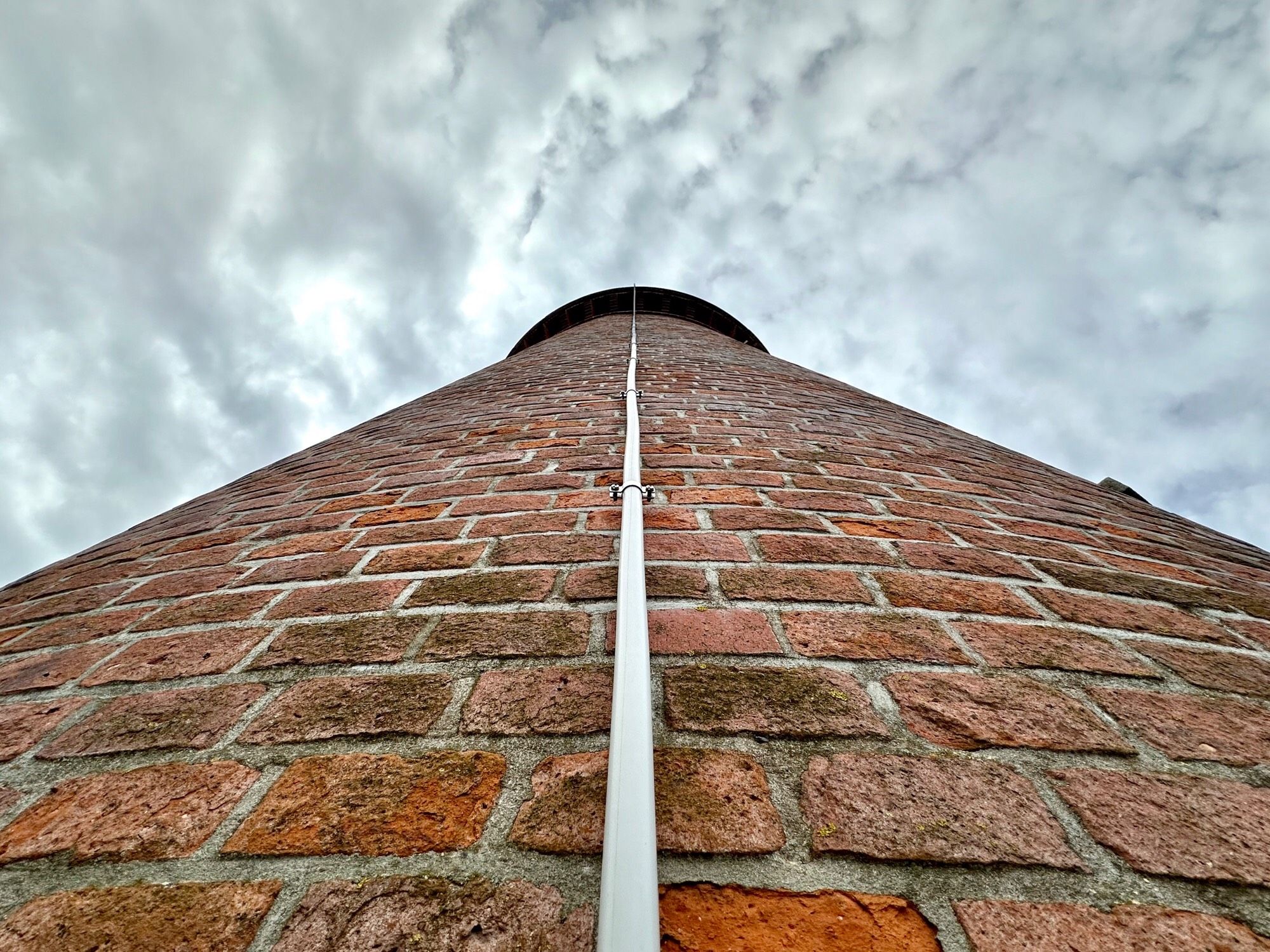 Farbfoto, Querformat, Zentralperspektive: Blick in den bewölkten Himmel an der Mauer eines Leuchtturms entlang. Der Turm ist rund und aus Backsteinen gemauert. In der Bildmitte verläuft senkrecht ein Blitzableiter.