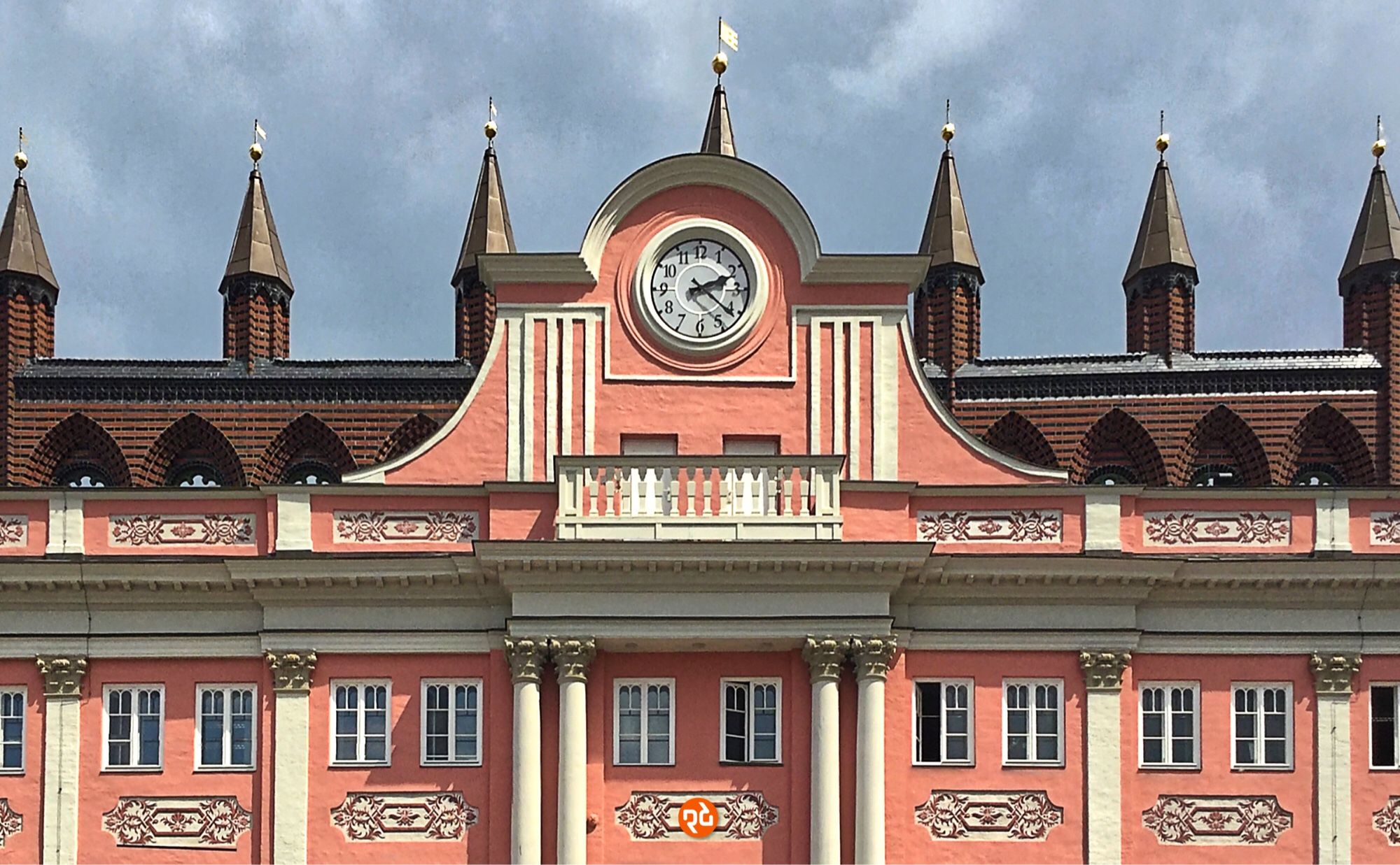 Farbfoto, Querformat, Ansicht der Fassade in Zentralperspektive. Die reich verzierte Fassade ist weiß und hellrot gestrichen. Die Rathausuhr in der Bildmitte zeigt 14:21 Uhr. Im Hintergrund erkennt man eine Backsteinwand mit sieben spitzen Türmchen. Darüber ein stark bewölkter grauer Himmel.