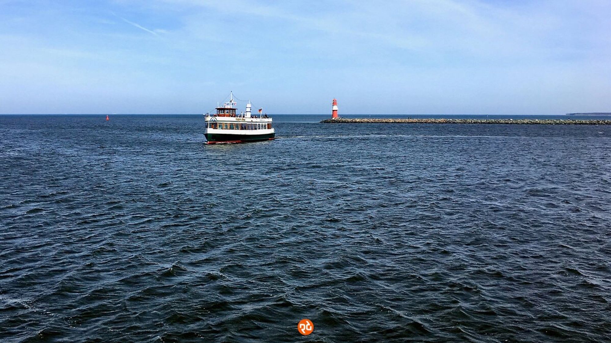 Farbfoto, Querformat, Panoramablick übers Meer, der Horizont teilt das Bild im oberen Drittel. Von rechts ragt eine flache Mole mit einem kleinen roten Leuchtturm an der Spitze bis fast in die Bildmitte. Links im Bild ein kleiner historischer Ausflugdampfer mit Kurs auf den Fotografen. Das Meer ist dunkel und leicht gekräuselt, überm Horizont ein blassblauer verschleierter Himmel.