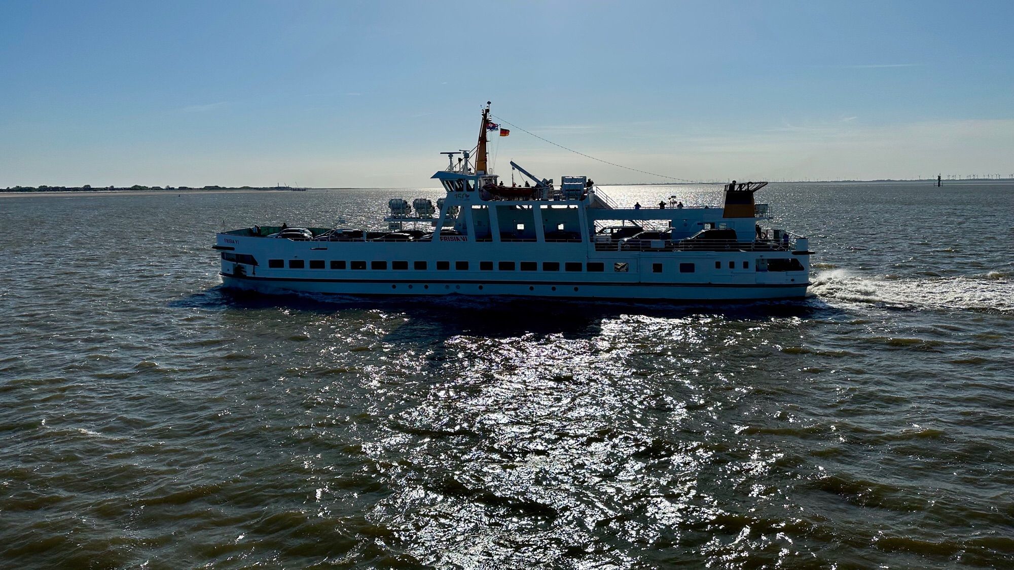 Farbfoto, Querformat: Die Fähre nach Norderney im Gegenlicht der aufgehenden Sonne. Das Schiff gleich einer dunklen Silhouette in der glitzernden Nordsee.