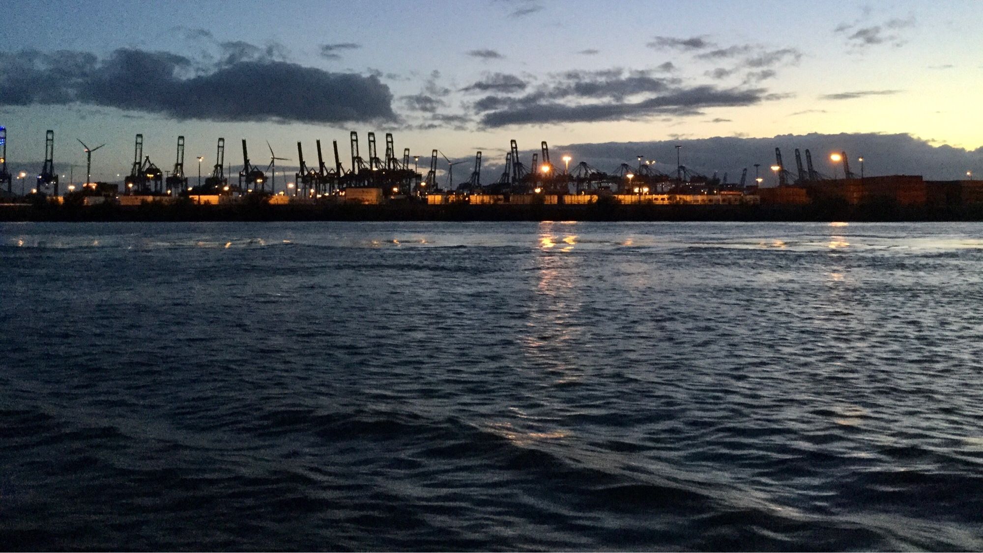 Farbfoto, Querformat, Panoramablick über die Elbe bei Sonnenuntergang: Im Vordergrund spiegelt sich der Abendhimmel im leicht gekräuselten Fluss. Am Horizont eine lange Reihe von beleuchteten Kränen. Darüber einige Wolken.