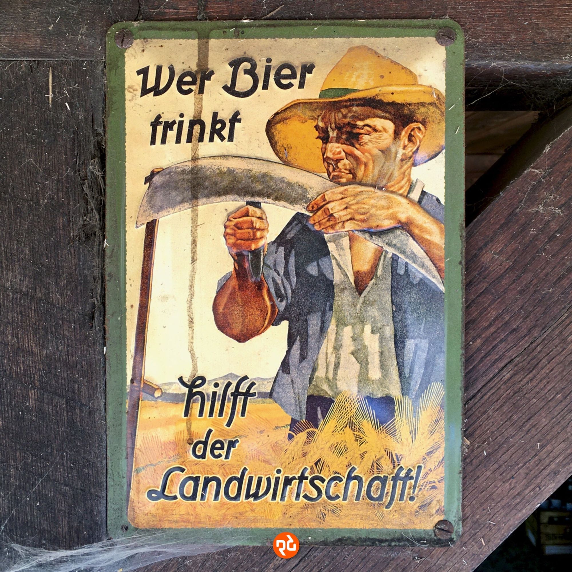 Farbfoto, quadratisch. Altes Reklameschild mit der Aufschrift »Wer Bier trinkt, hilft der Landwirtschaft!« Dazu ist die farbige Zeichnung eines Getreidebauers abgebildet, der gerade seine Sense schärft. Das Schild ist an einem dunkelbraunen Balken befestigt.