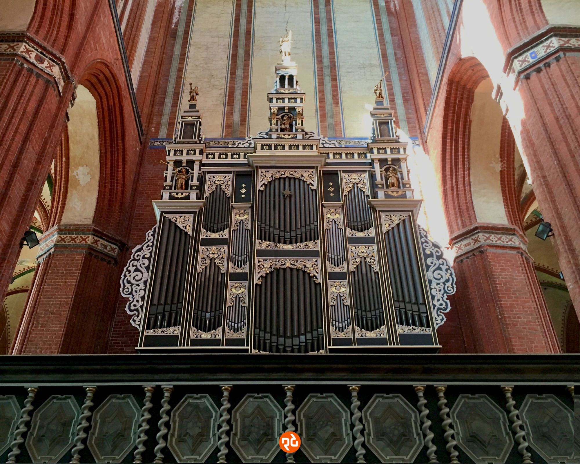 Farbfoto, Querformat: Ansicht des Klangkörpers der Orgel schräg von unten in Zentralperspektive. Die schwarz gefärbten Orgelpfeifen stecken in einem mit Reliefs und Engelsstatuetten reich verzierten Gehäuse. Beiderseits der Orgel erkennt man aufstrebende Säulen aus Backstein. Im Vordergrund eine dunkle, mit kleinen Säulen und Schilden verzierte Balustrade.