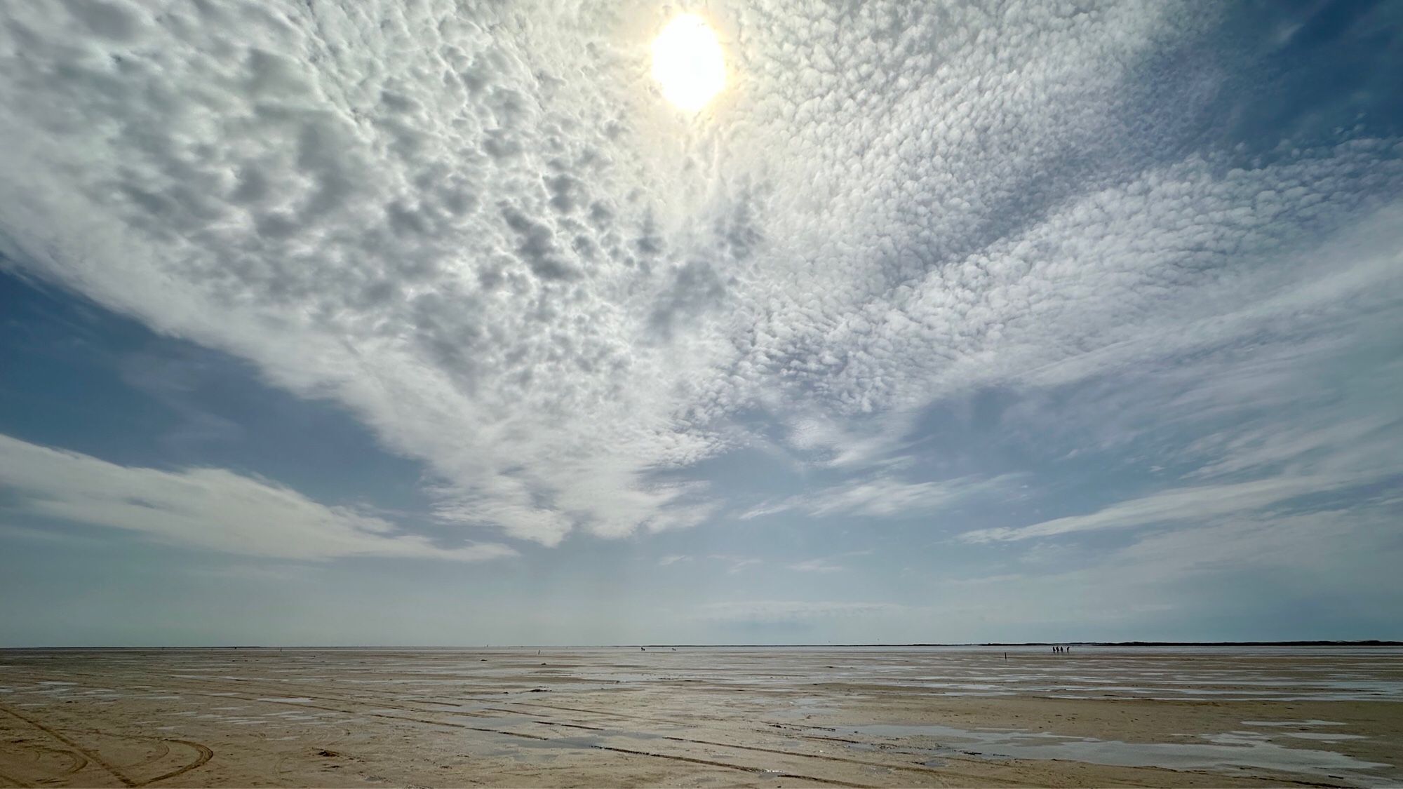 Farbfoto, Querformat, Panoramablick übers Wattenmeer bei Ebbe. Im Vordergrund der Meeresboden mit letzten Pfützen und Prielen, über dem Horizont ein fächerförmiges Wolkengebilde. Am oberen Bildrand in der Mitte behauptet sich die Mittagssonne gegen die Wolken. Am Horizont rechts winzig klein eine Gruppe von fünf Wanderern.