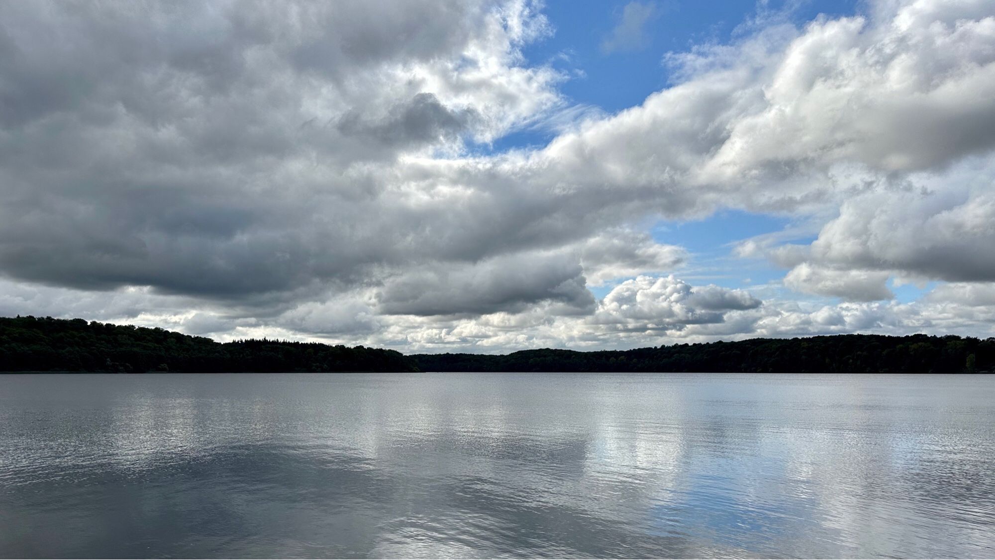 Farbfoto, Querformat, Panoramablick über den See: Eine große graue Wolkenbank und ein kleiner Rest vom blauen Himmel spiegeln sich in der leicht gekräuselten Wasserfläche. Am Horizont erkennt man das dicht bewaldete gegenüberliegende Ufer. Der Horizont teilt das Bild im unteren Drittel.