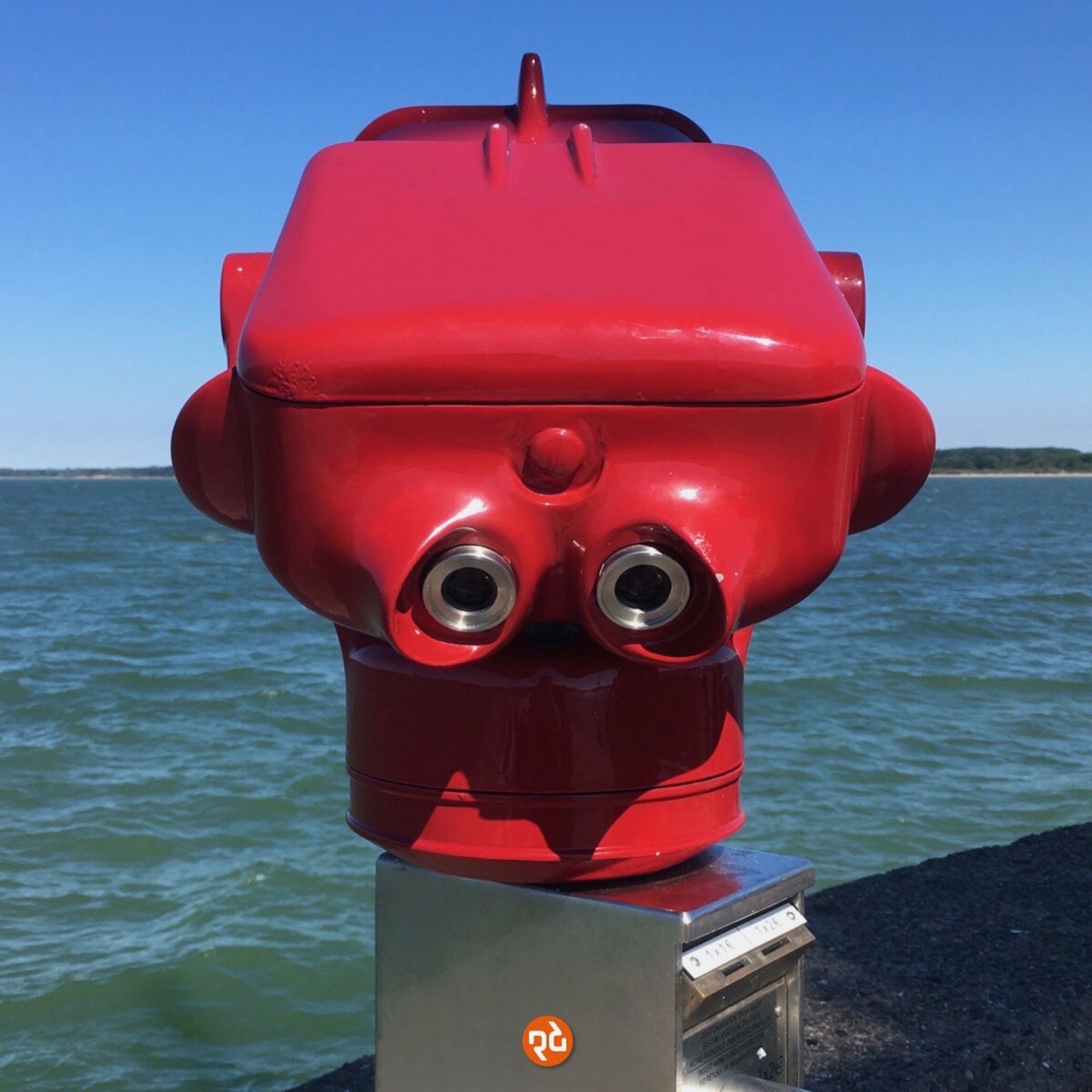 Farbfoto, quadratisch: Traurig blickendes zweiäugiges rotes Fernrohr vor blauem Himmel an der Promenade von Travemünde. Im Hintergrund leicht gekräuseltes dunkles Wasser.