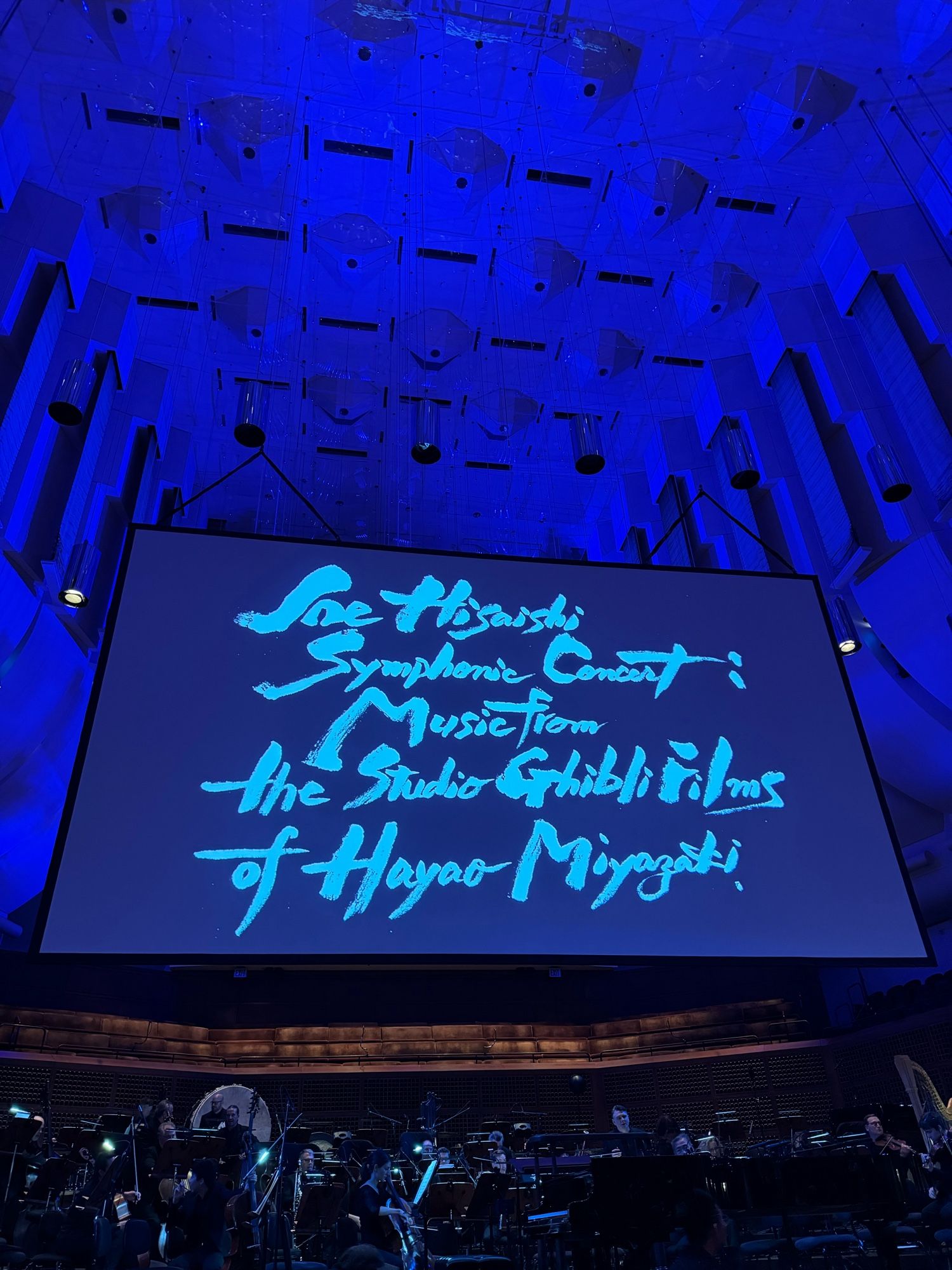 A screen over the stage of Davies Symphony Hall in San Francisco has a title card that reads, “Joe Hisaishi Symphonic Concert: Music from the Studio Ghibli Films of Hayao Miyazaki”