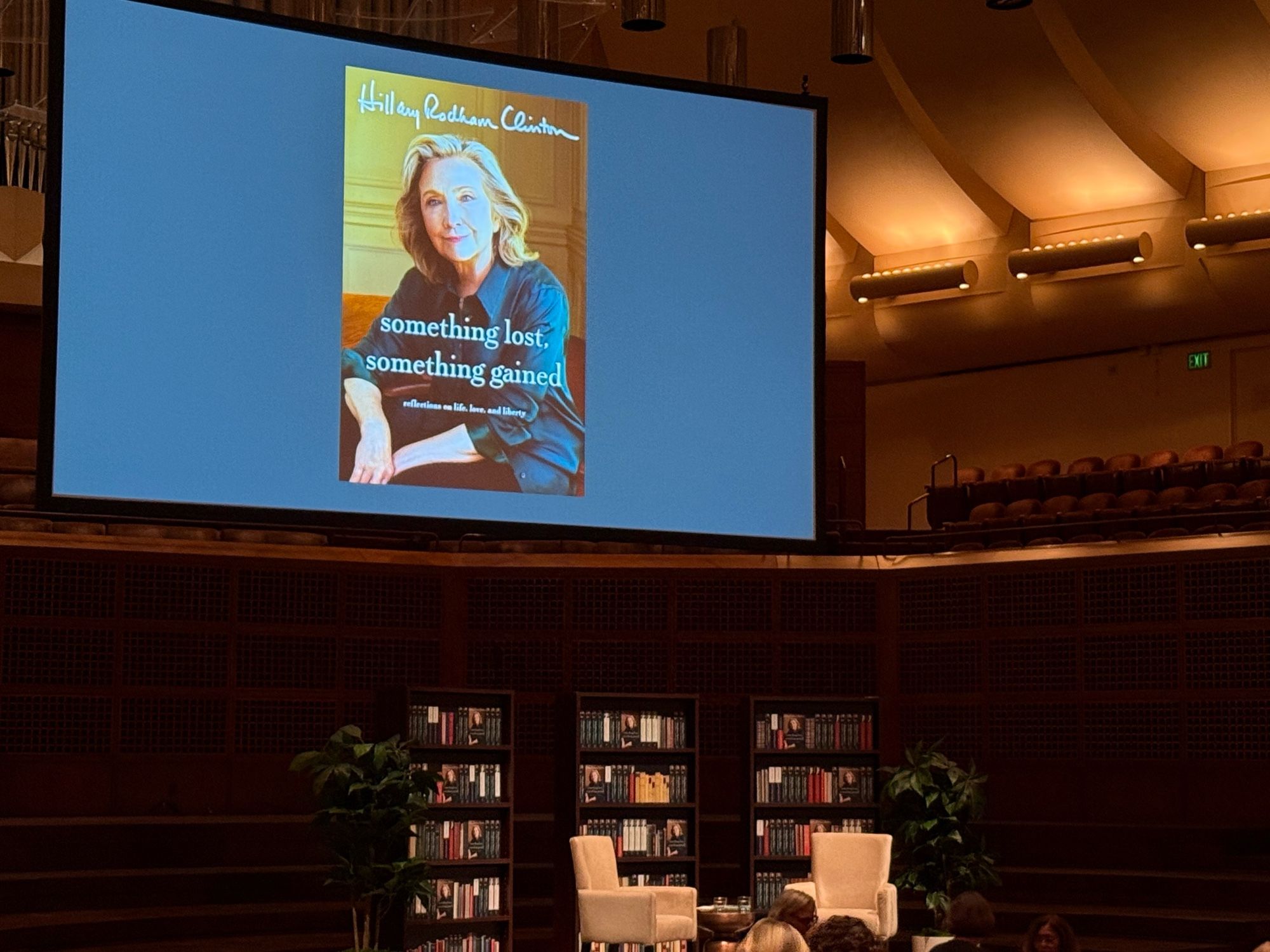 The stage of Davies Symphony Hall in San Francisco is set with a pair of armchairs, a small table between them, three full bookcases behind them, and pair of potted plants flanking the bookcases, and a large screen hanging above it all. The screen shows the cover of Hillary Clinton’s latest book, “Something Lost, Something Gained”. The bookcases also feature some copies of that book.