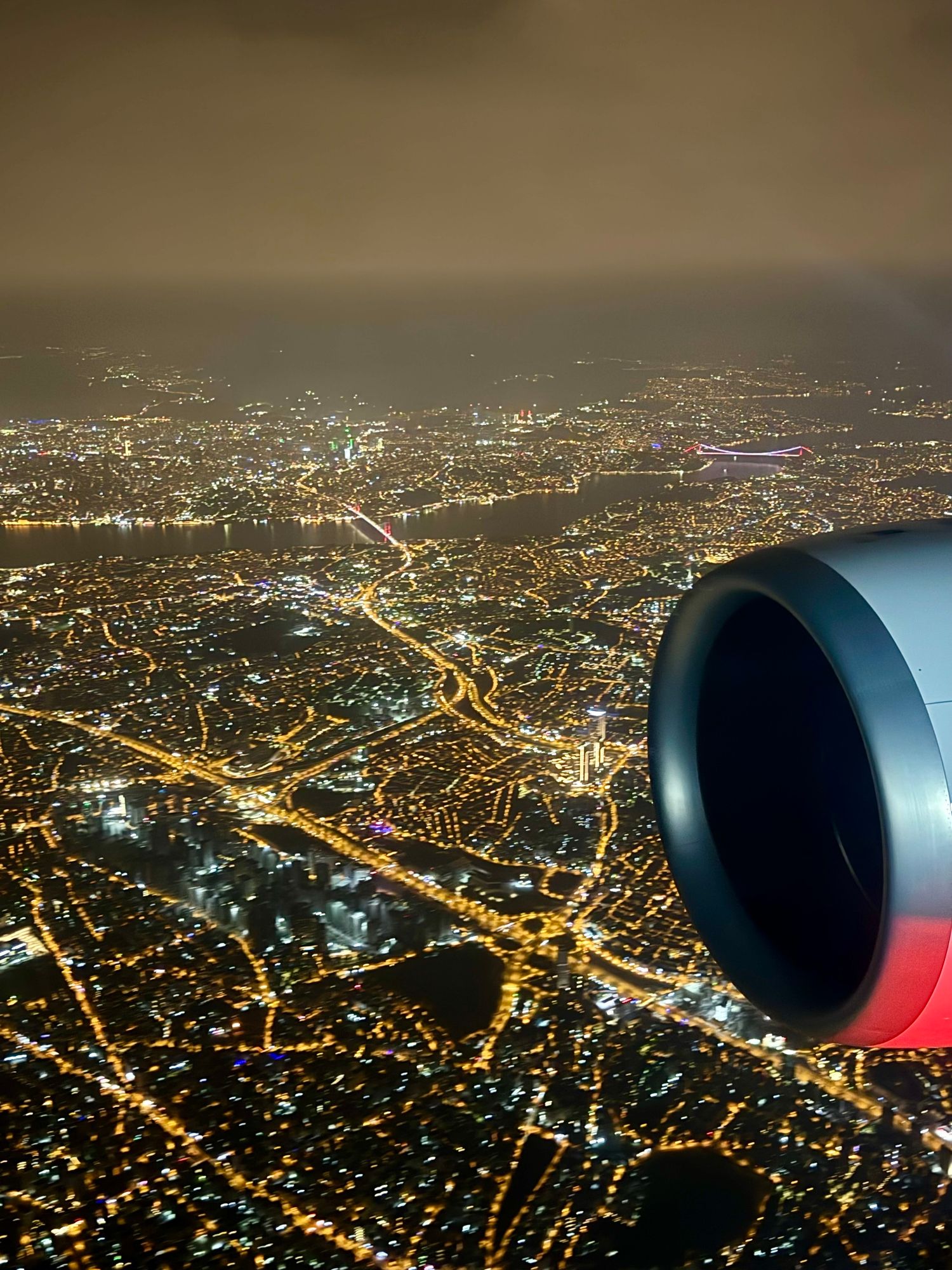 Photo out of starboard window of aircraft, at night, showing the city of Istanbul below, with the front on an aircraft engine in the foreground 