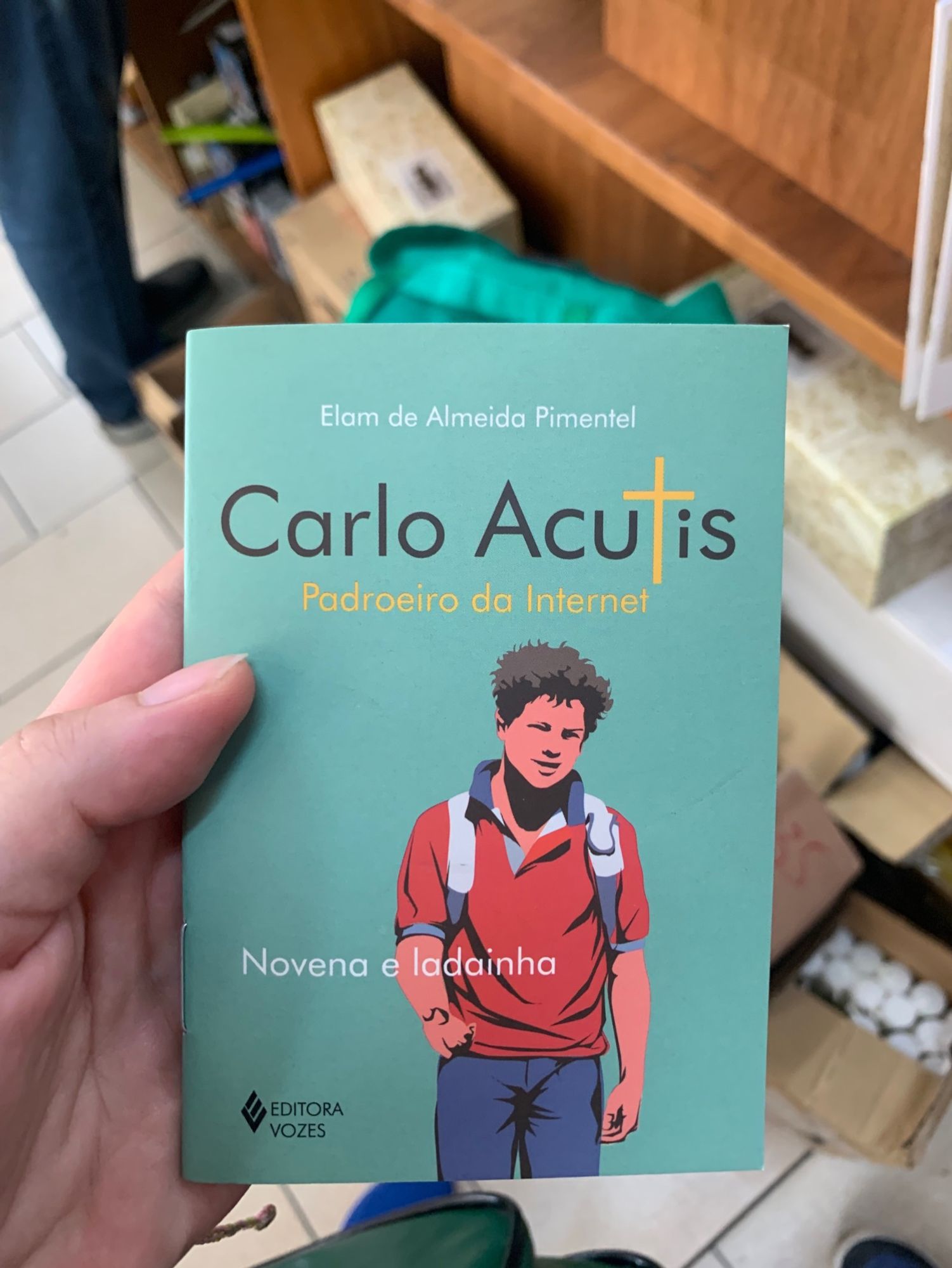 Livro de novena azul piscina com a imagem de um jovem de cabelo castanho, pele branca, com calça jeans, blusa vermelha e mochila nas costas, representando são carlo acutis, padroeiro da internet