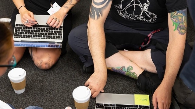 Kaz and Simon sitting cross-legged on the floor at a OneTeamGov event some years back; macbooks, coffee cups, black clothes and tattoos are the main features! I think the original photo credit is Sam Villis