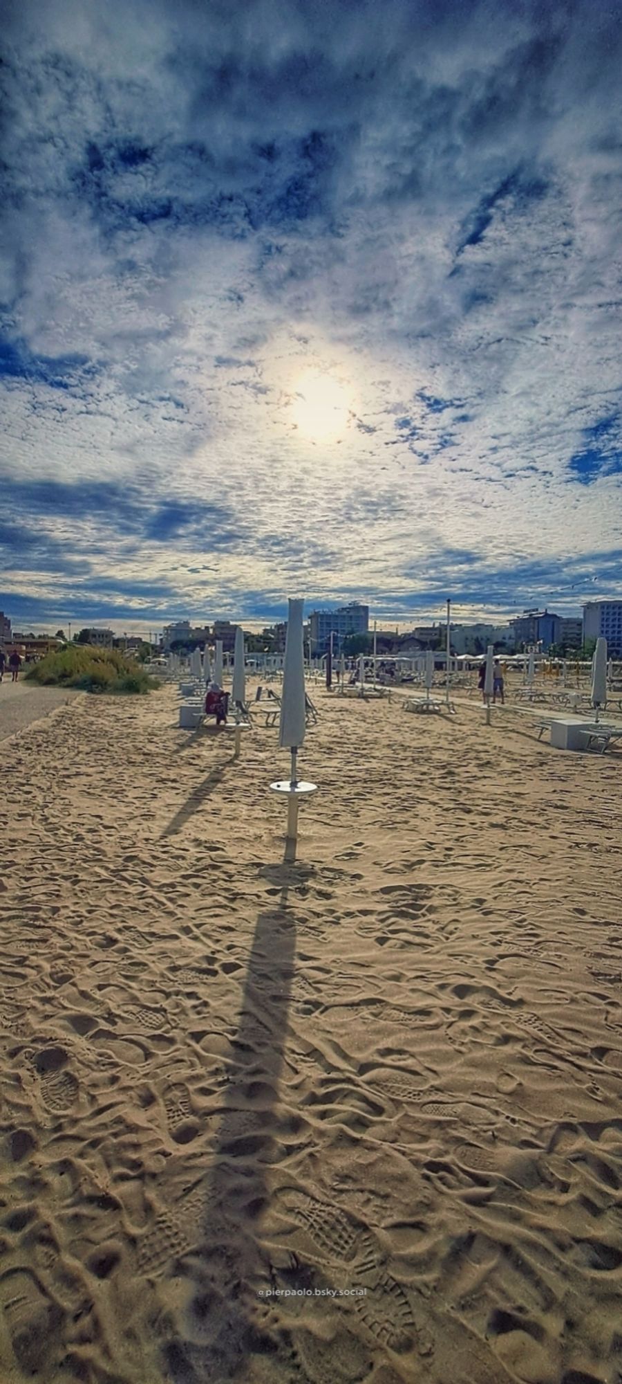 La spiaggia di fronte a Piazza J.F.Kennedy a Rimini nel tardo pomeriggio. Il sole, anche se nascosto dalle nubi, riesce ad illuminare la spiaggia semi deserta con gli ombrelloni chiusi la cui ombre si allungano sulla sabbia