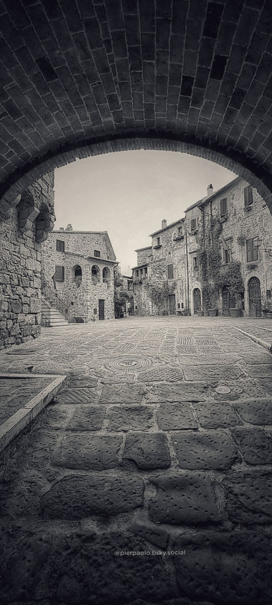 Attraverso un arco a sesto ribassato di mattoni si scorge l'abitato medioevale di Montemerano, un piccolo villaggio nel comune di Manciano (Grosseto). Le case in pietra a vista sono adornate da piante rampicanti. Anche la strada è  lastricata di pietre.