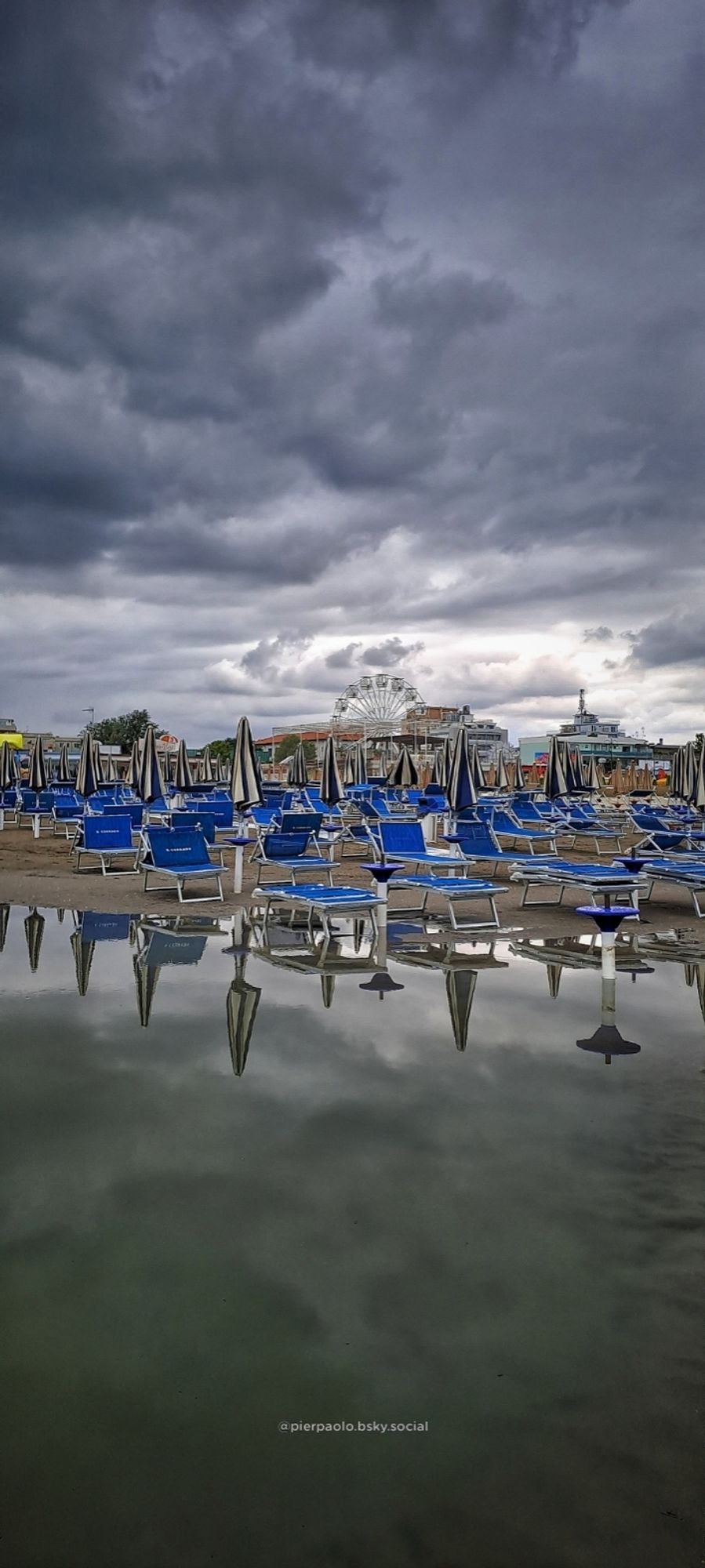 Ombrelloni e i lettini sono riflessi nel mare dopo che una mareggiata ha eroso una parte della spiaggia. Il cielo è  coperto di nubi grigie che minacciano pioggia.