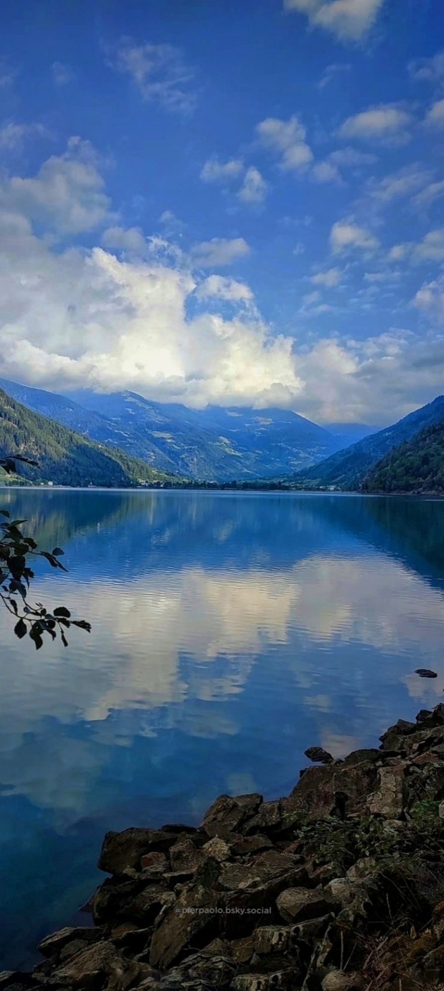 Il Lago di Poschiavo (Svizzera) fotografato dalla località Miralago. Le montagne che lo circondano e il cielo azzurro coperto di nuvole bianche si riflettono sul lago.