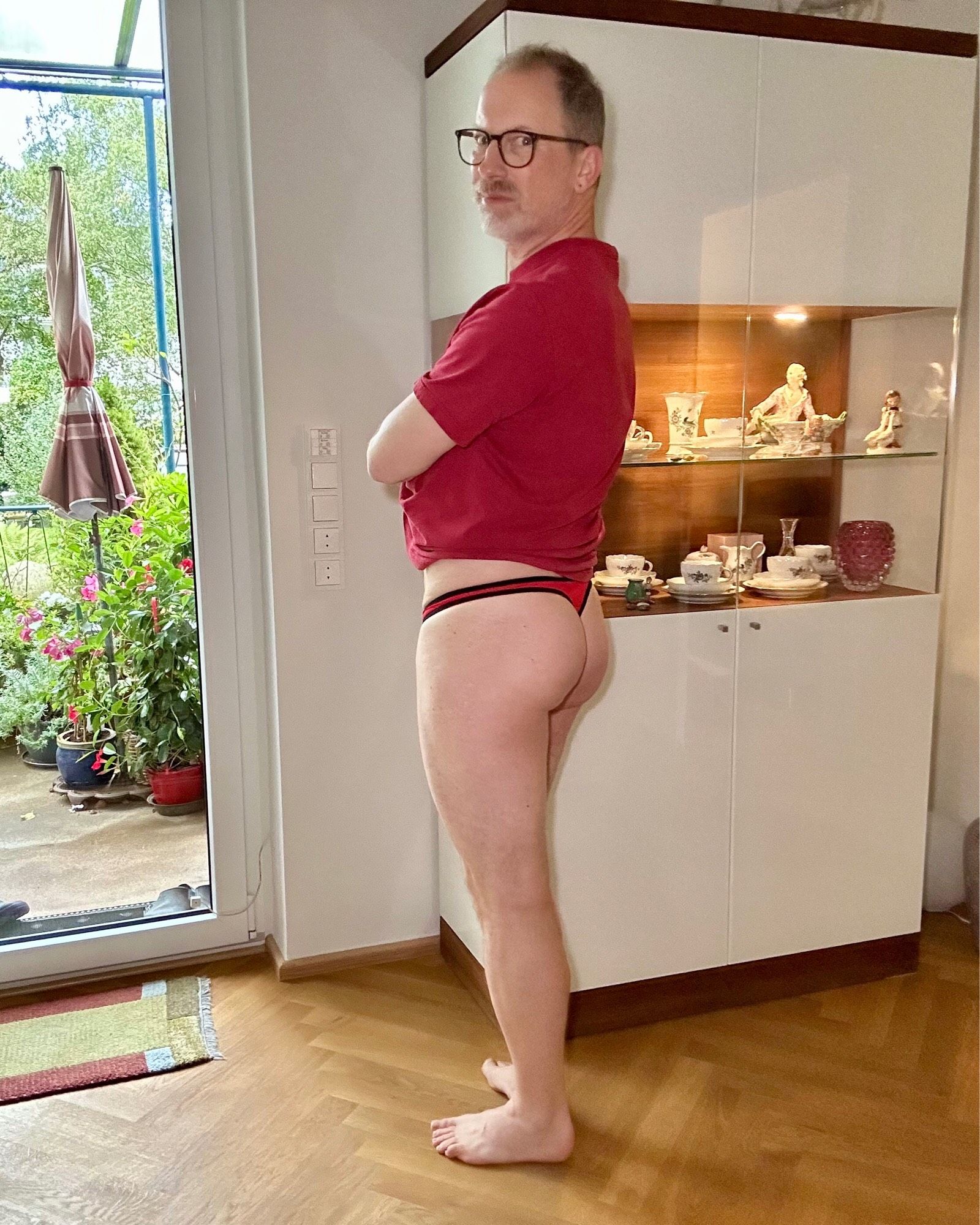 Facing away from the camera but face turned around, wearing a red manthong and a matching tee. Shirt is slid up to display the thong back and my bare bum. Face not looking happy