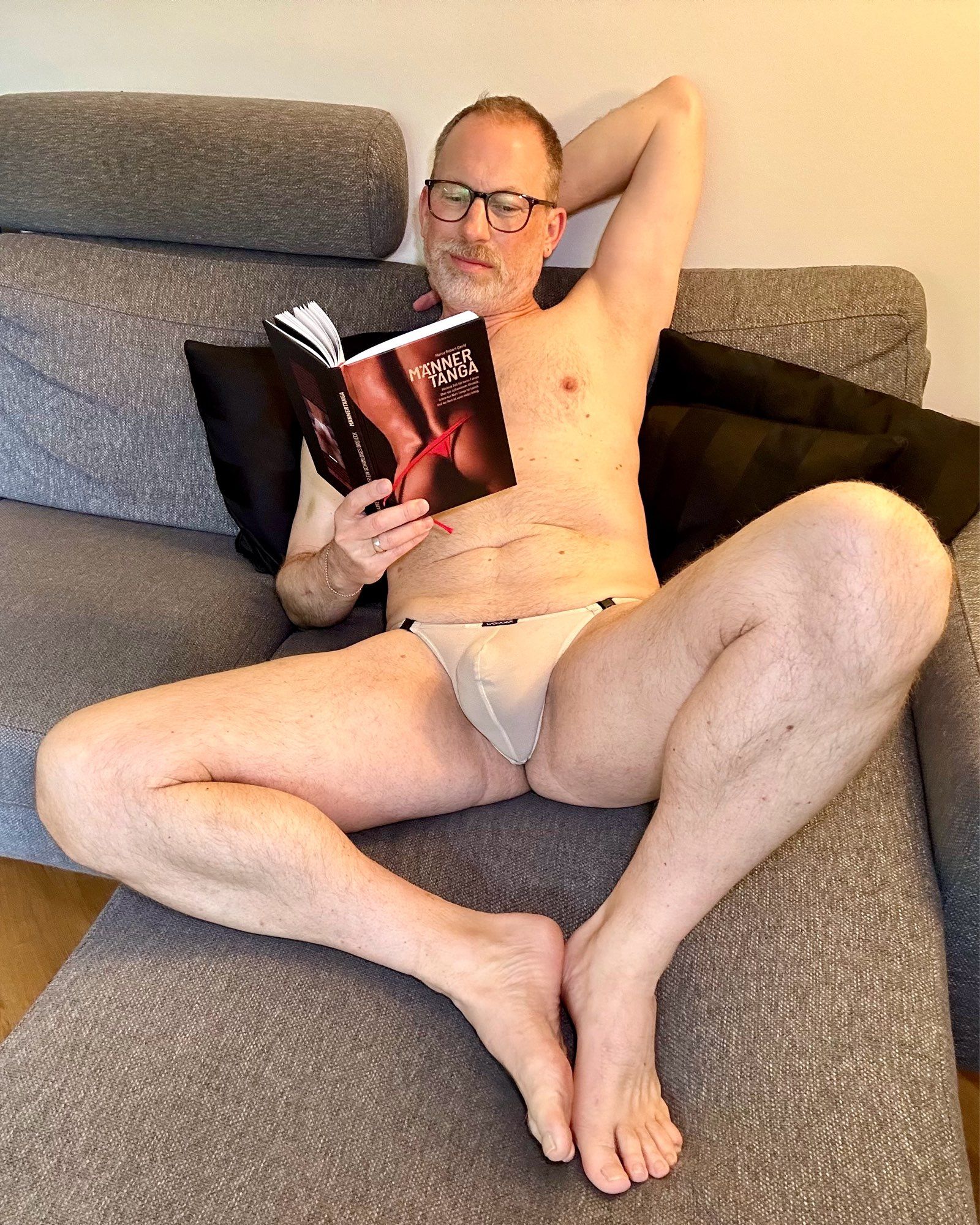 Layed back on the sofa, one arm behind my head, still only wearing a thong in nude, reading the book „Männertanga“ (Manthong)