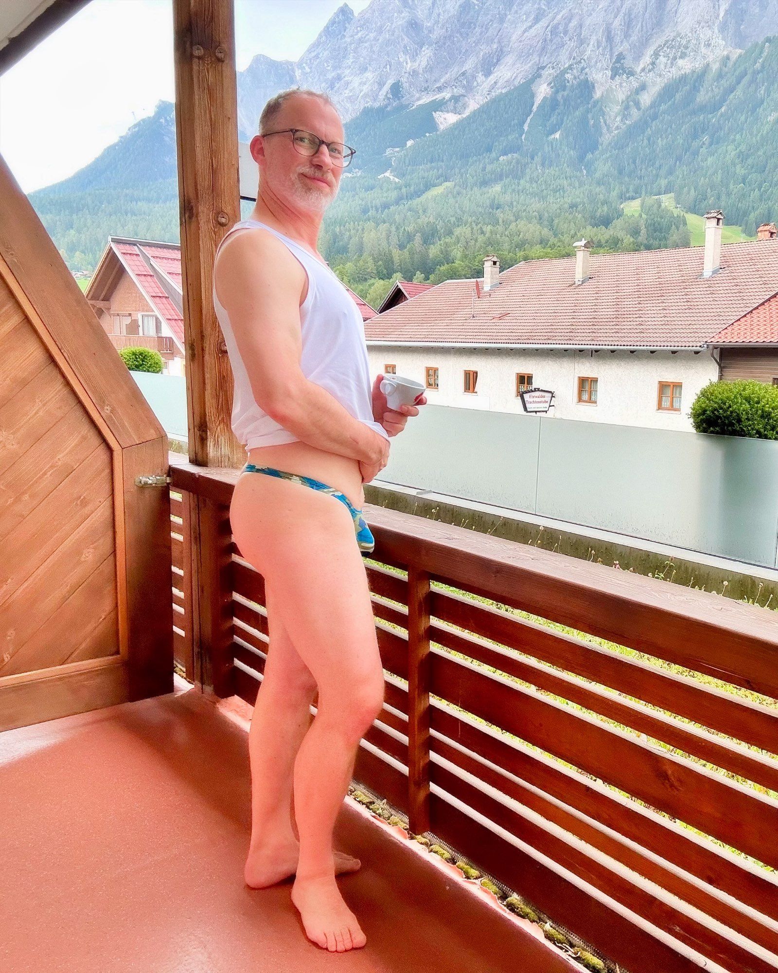 Standing on a balcony with houses and mountains in the background, holding a cup of coffee on one hand, only wearing a blue camouflage thong and a white muscle shirt