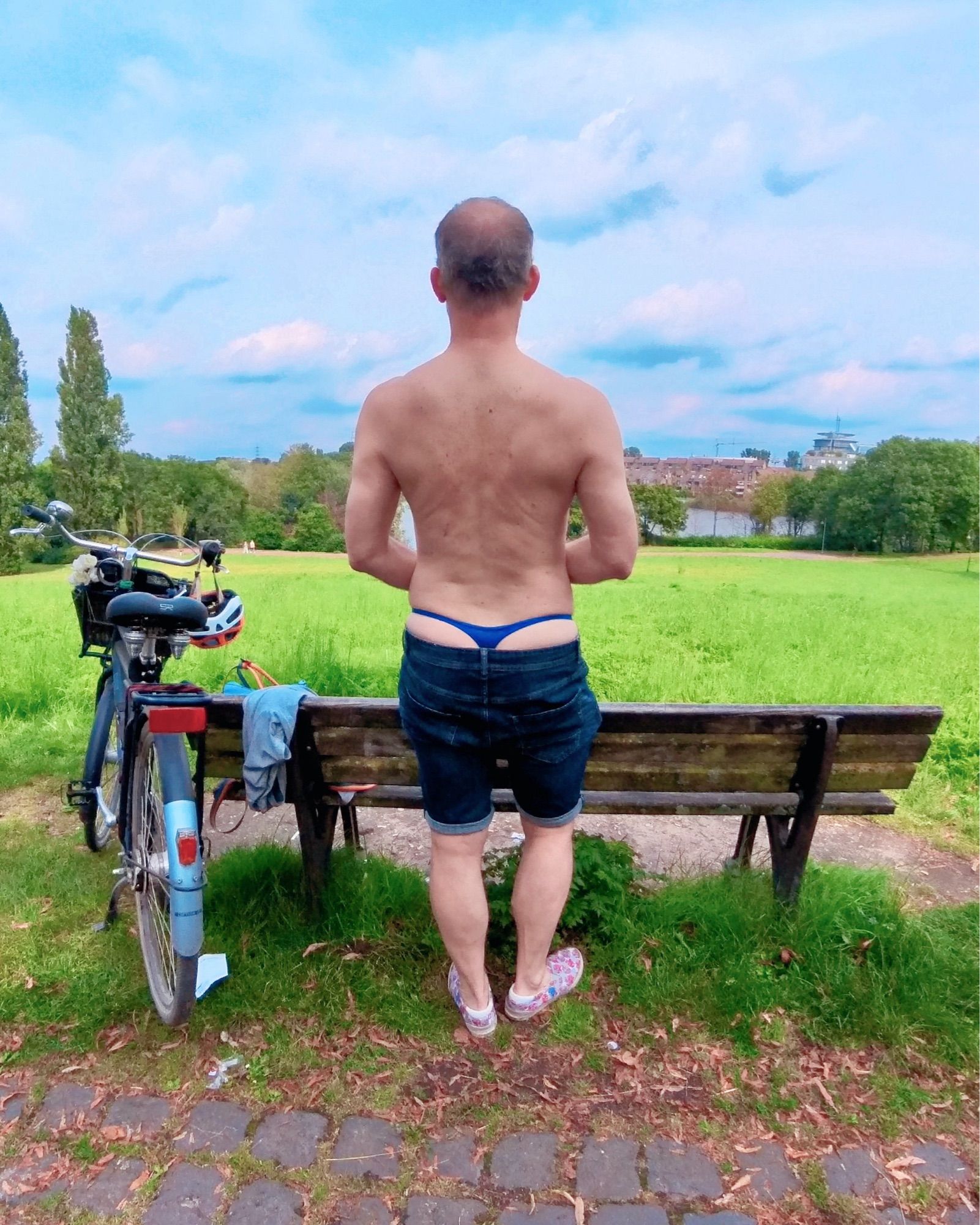 Standing in front of a park bench, facing away from the camera, wearing only jeans shorts and shoes, whale tale of my blue Wojoer thong is visible while I enjoy the view of the scenery. Next to the bench my bicycle is parked