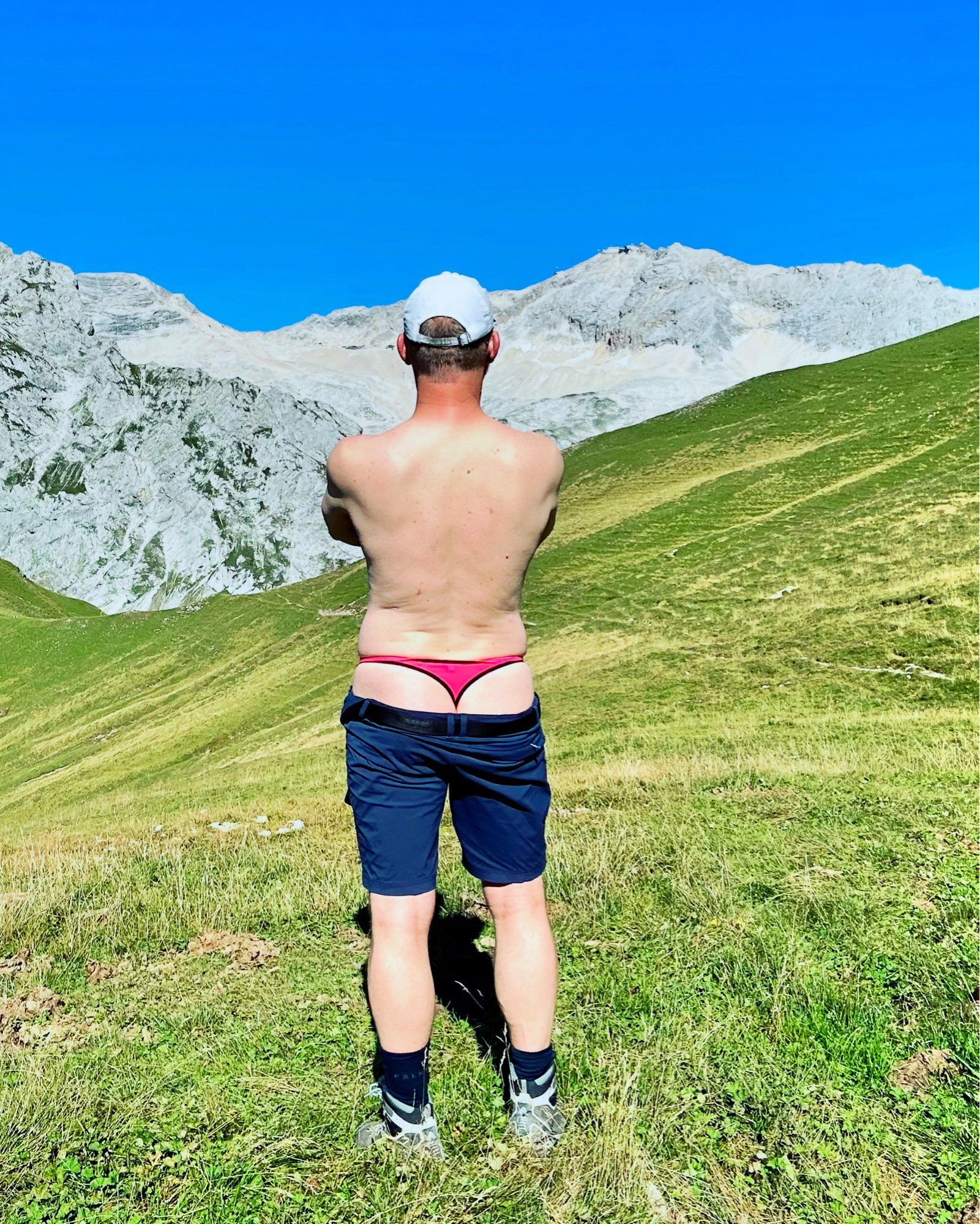 Standing on a lawn facing away from the camera, showing my manthong underneath my hiking shorts, the Zugspitze (Germanys highest mountain) in the background