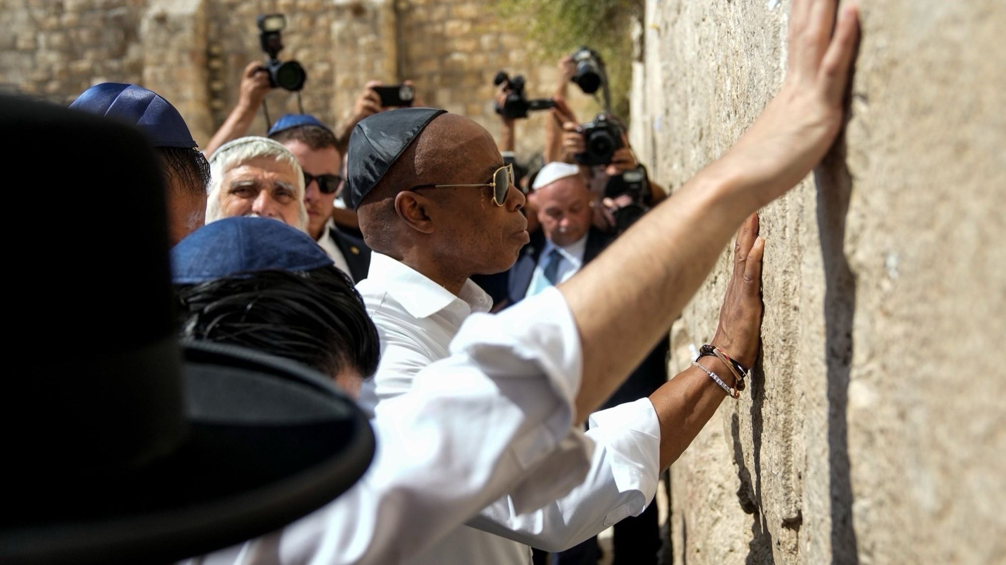 Eric Adams in Jerusalem, wearing a kippah but also sunglasses and a lettered bracelet. He is touching the Western Wall