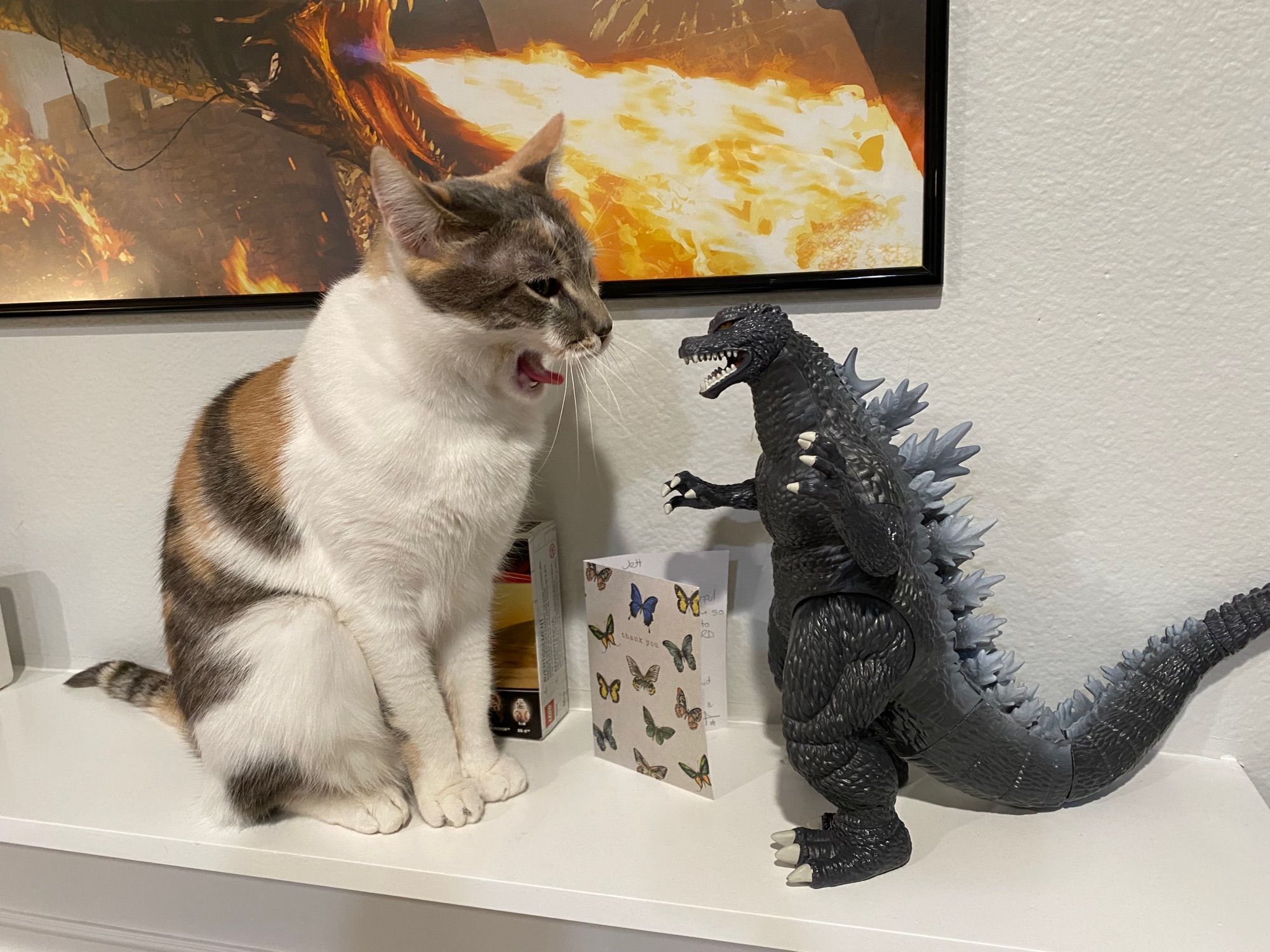 Yara (a mostly white calico cat) sitting on a white shelf next to a large, plastic figure of Godzilla. She is mid-yawn with her tongue sticking out, and looks not entirely dissimilar to Godzilla (whose mouth is also open). Art of Balerion the Black Dread, an enormous dragon from “A Song of Ice and Fire,” can be seen on a white wall behind them. Balerion’s mouth is also open, but for breathing fire!