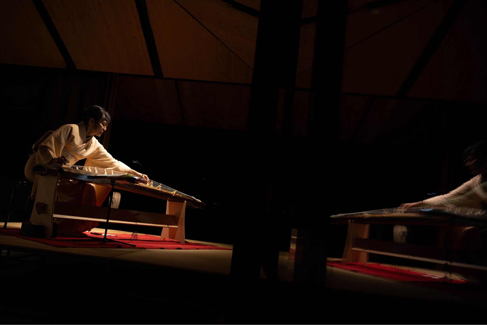 A dimly lit stage with a person in traditional Japanese clothing sitting on the floor, playing a koto (a long Japanese zither). The performer is illuminated by a spotlight, creating a dramatic contrast against the dark background. A reflection of the musician is visible, giving the initial impression of two players. Red cloth is visible beneath the instrument, adding a warm accent to the scene.​​​​​​​​​​​​​​​​